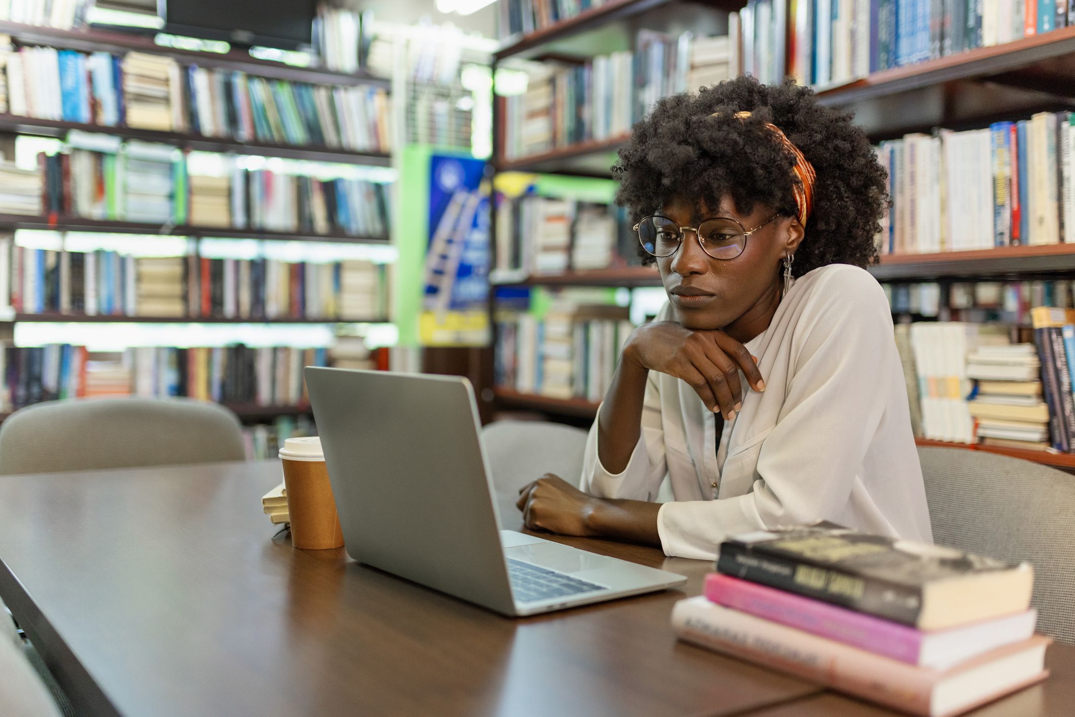 A researcher working on a laptop at a table surrounded by books in a library.