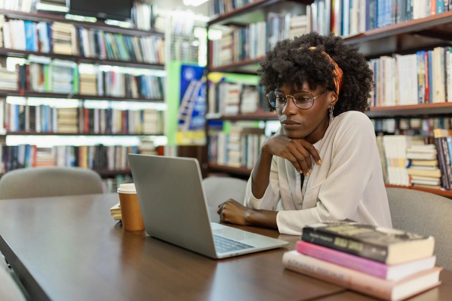 A researcher working on a laptop at a table surrounded by books in a library.