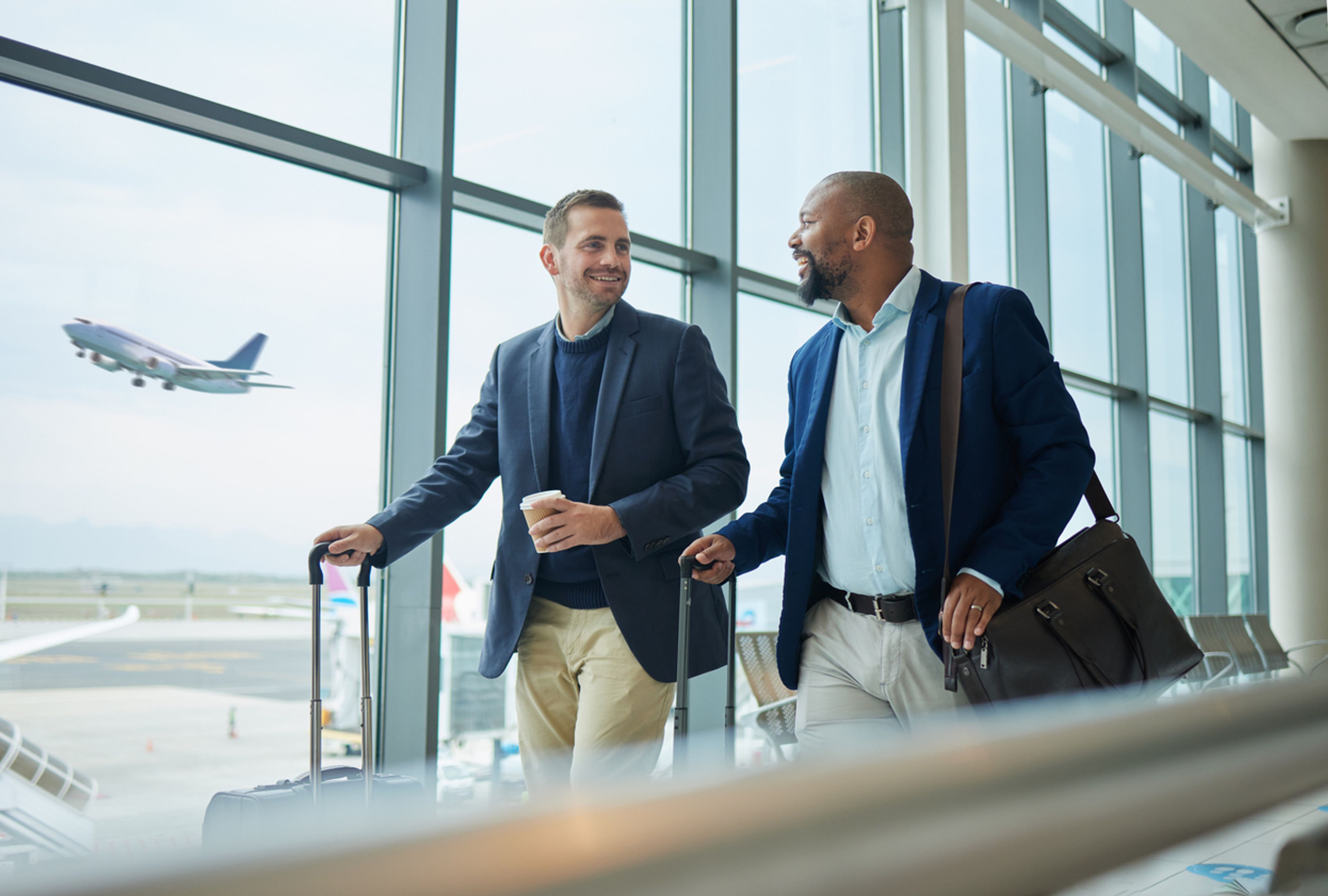 Two businessmen with luggage walk past an airport window as an airplane takes off in the background.