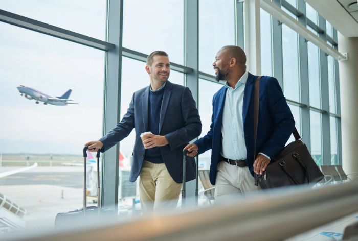 Two businessmen with luggage walk past an airport window as an airplane takes off in the background.