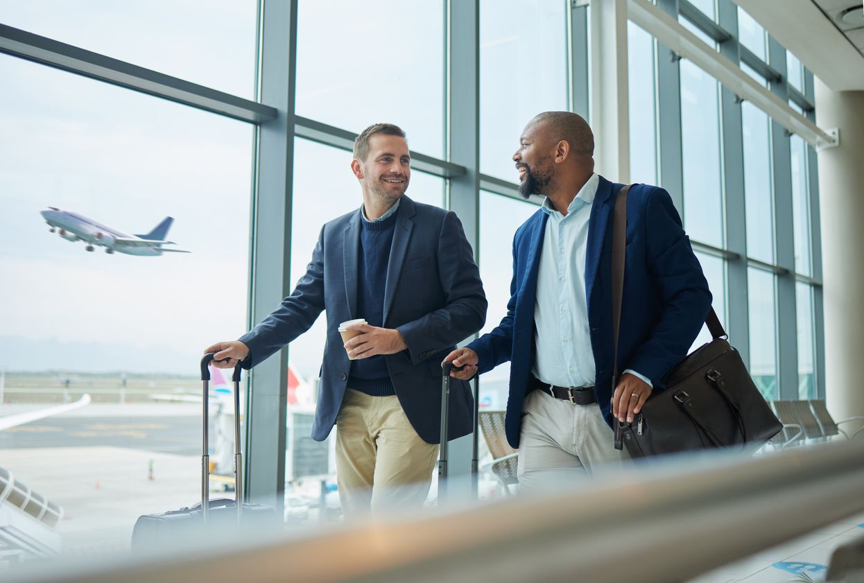 Two businessmen with luggage walk past an airport window as an airplane takes off in the background.