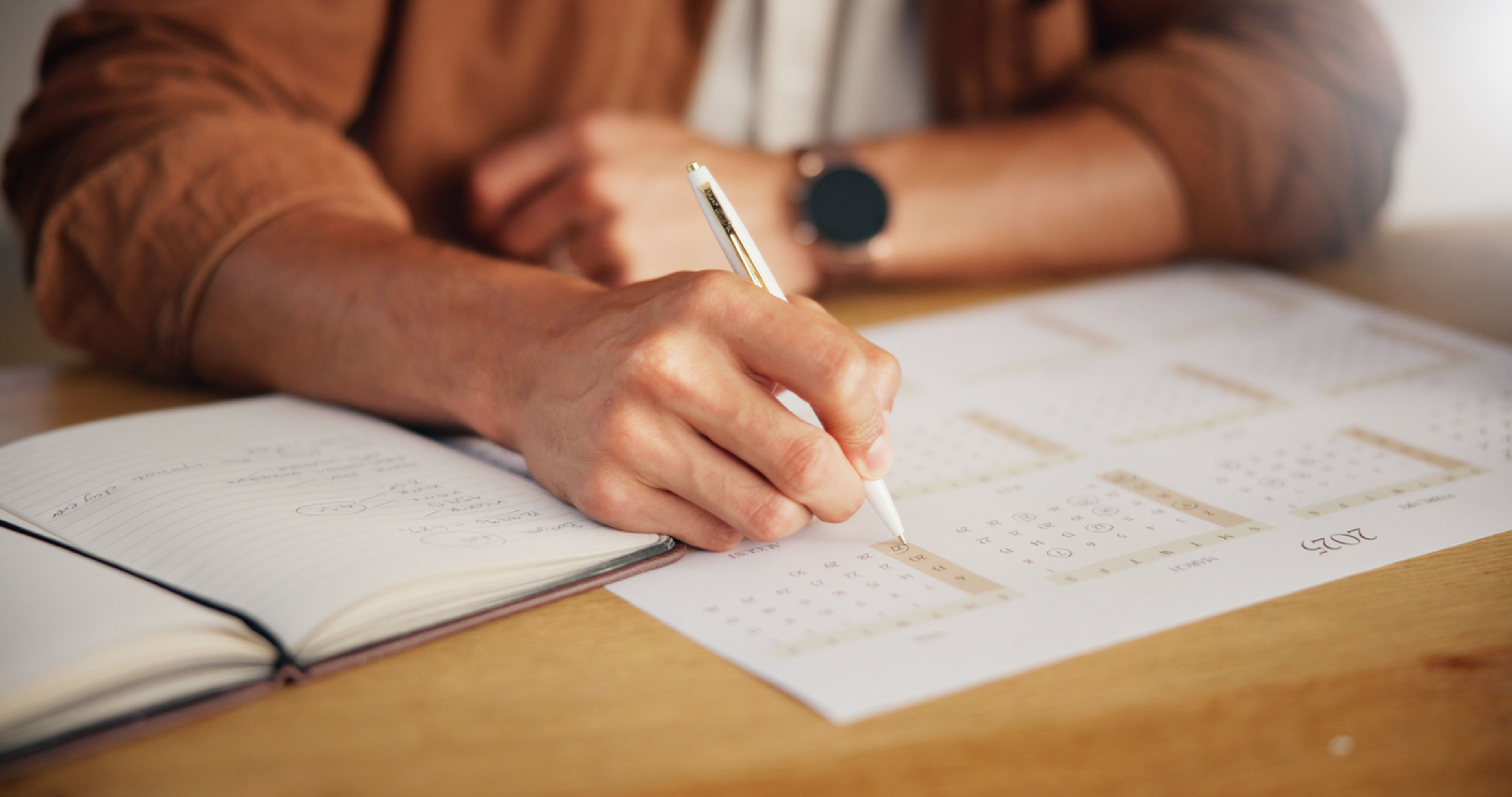 Closeup of a person's hand marking a date on a calendar next to an open planner.