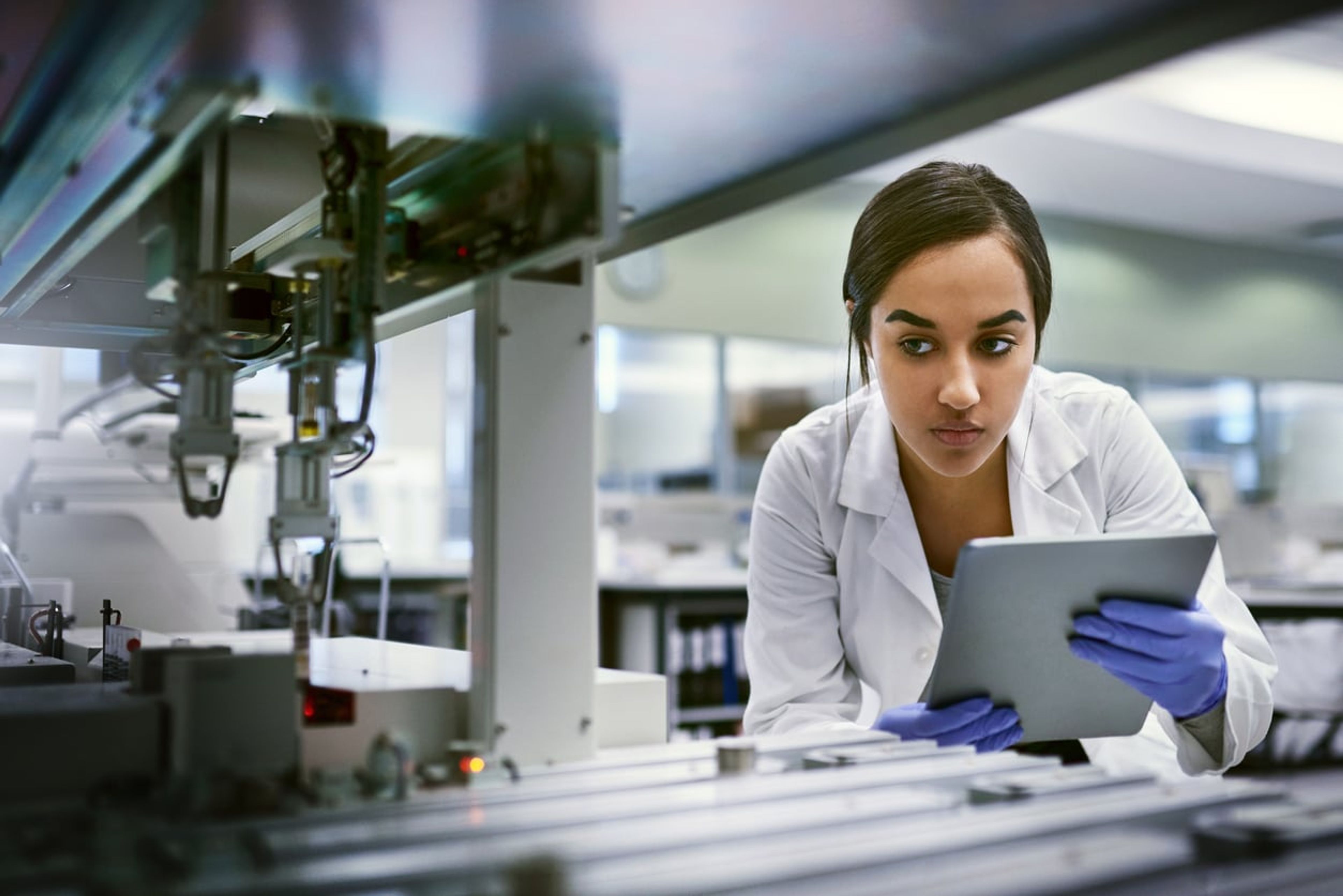 A young woman scientist takes notes on a digital tablet while watching an experiment in a laboratory.