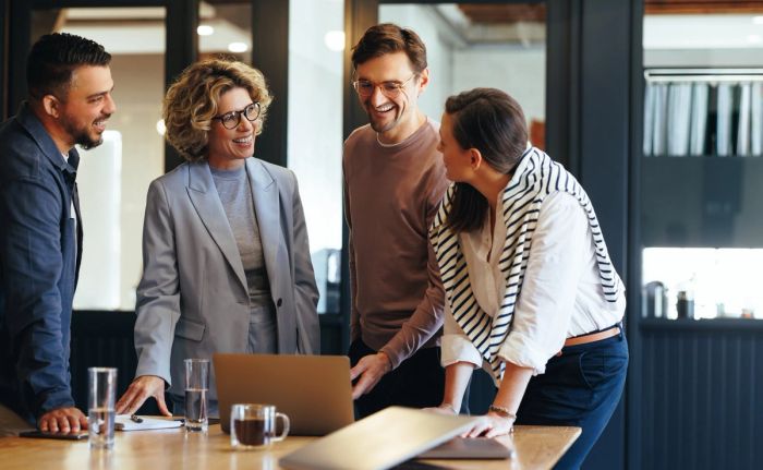A group of executive business people stand around a table with a laptop and talk during a meeting.