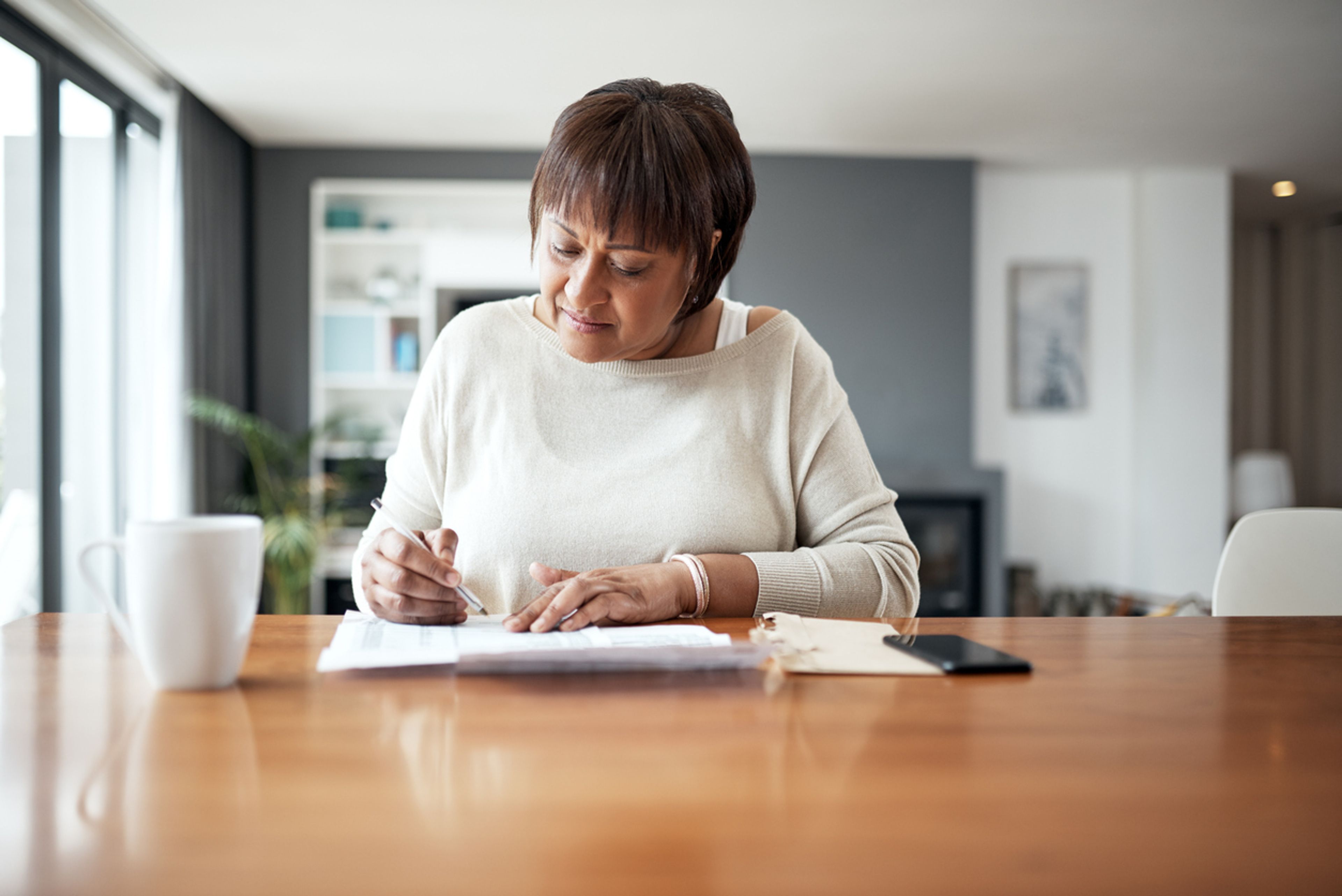 A mature woman in a white sweater sits at a table filling out a paper Form I-601A.