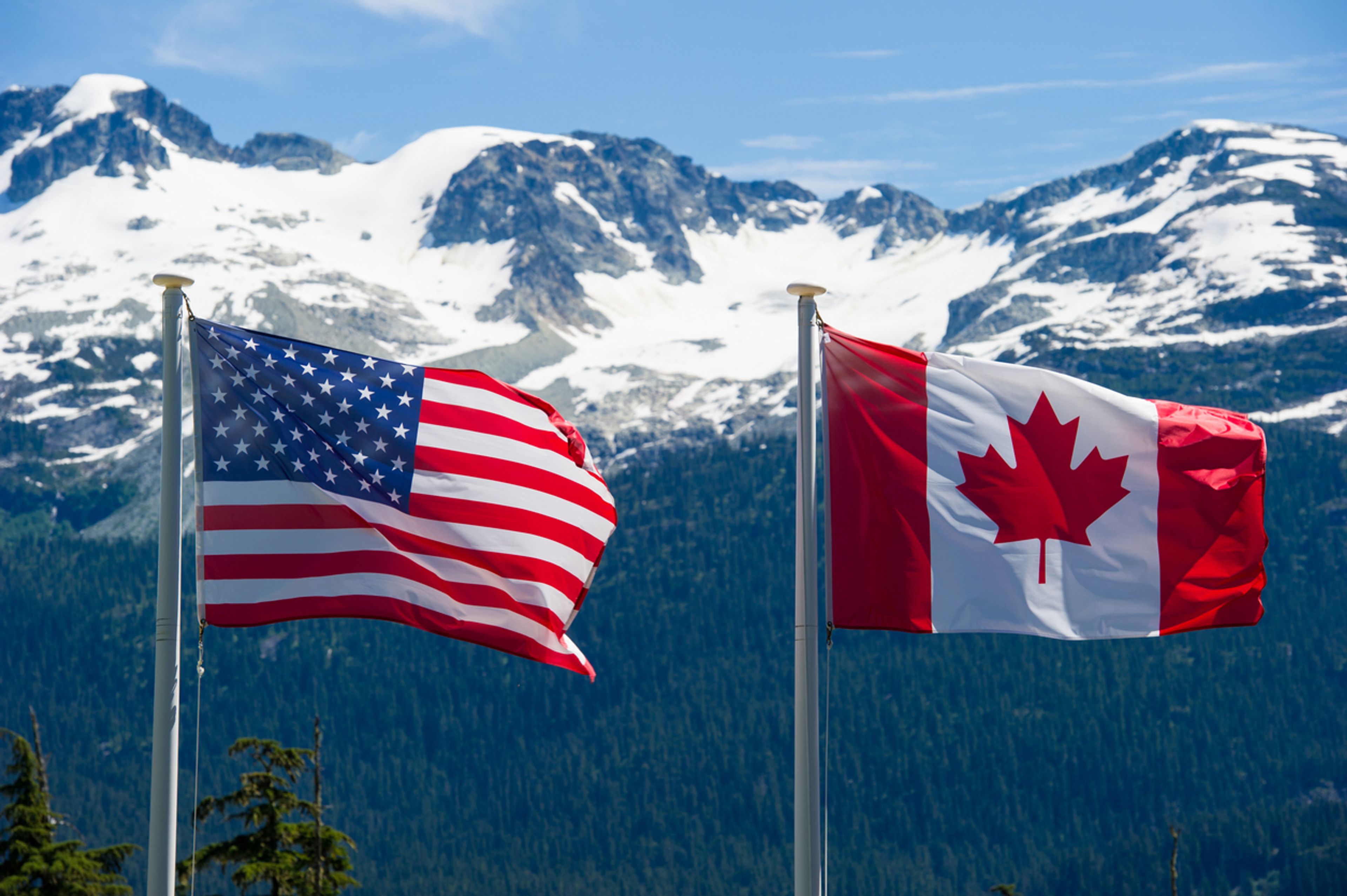 Canadian and American flags flying together in front of a mountain range