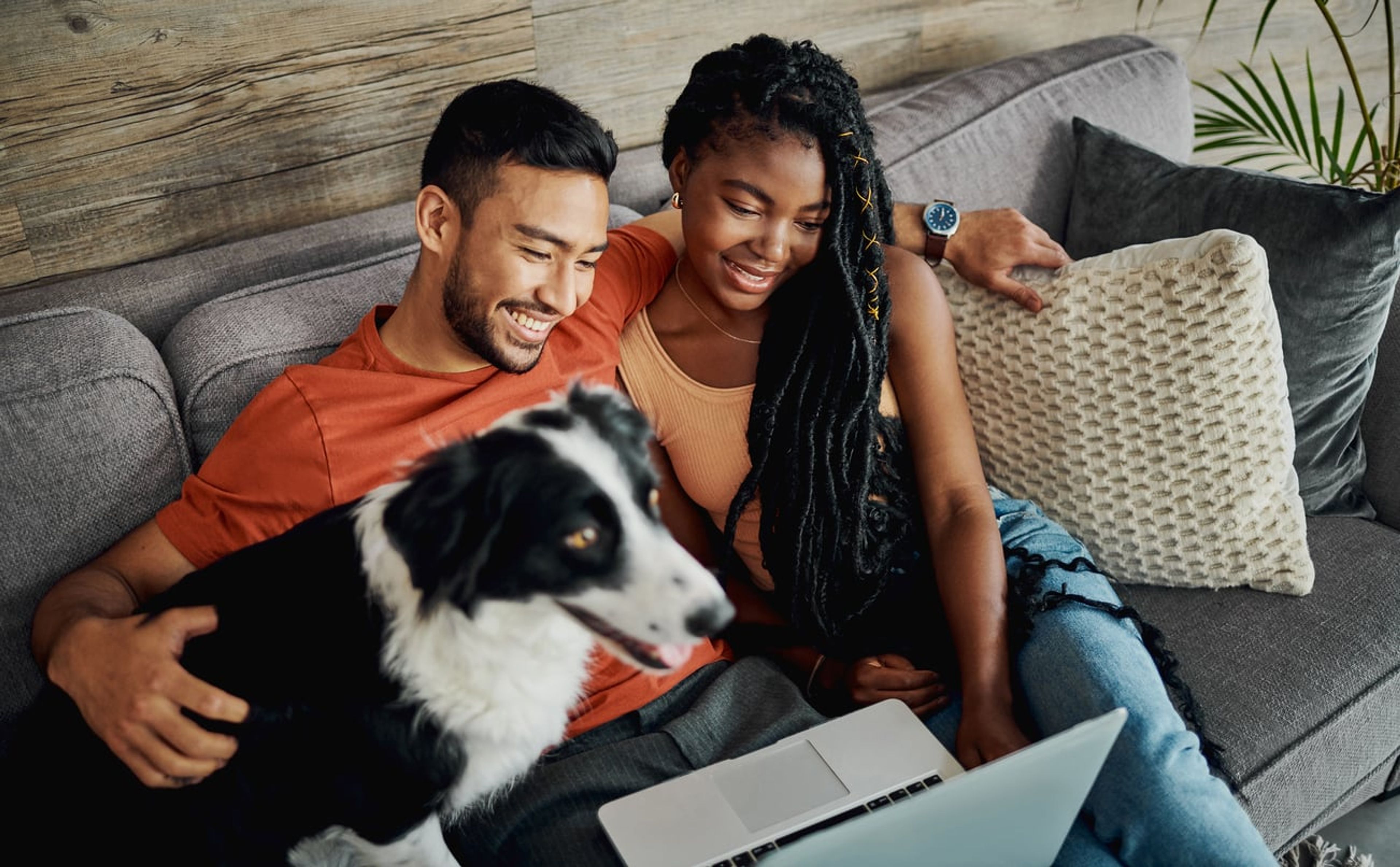 A married man and woman look at a laptop while sitting on a couch with their dog and smiling.