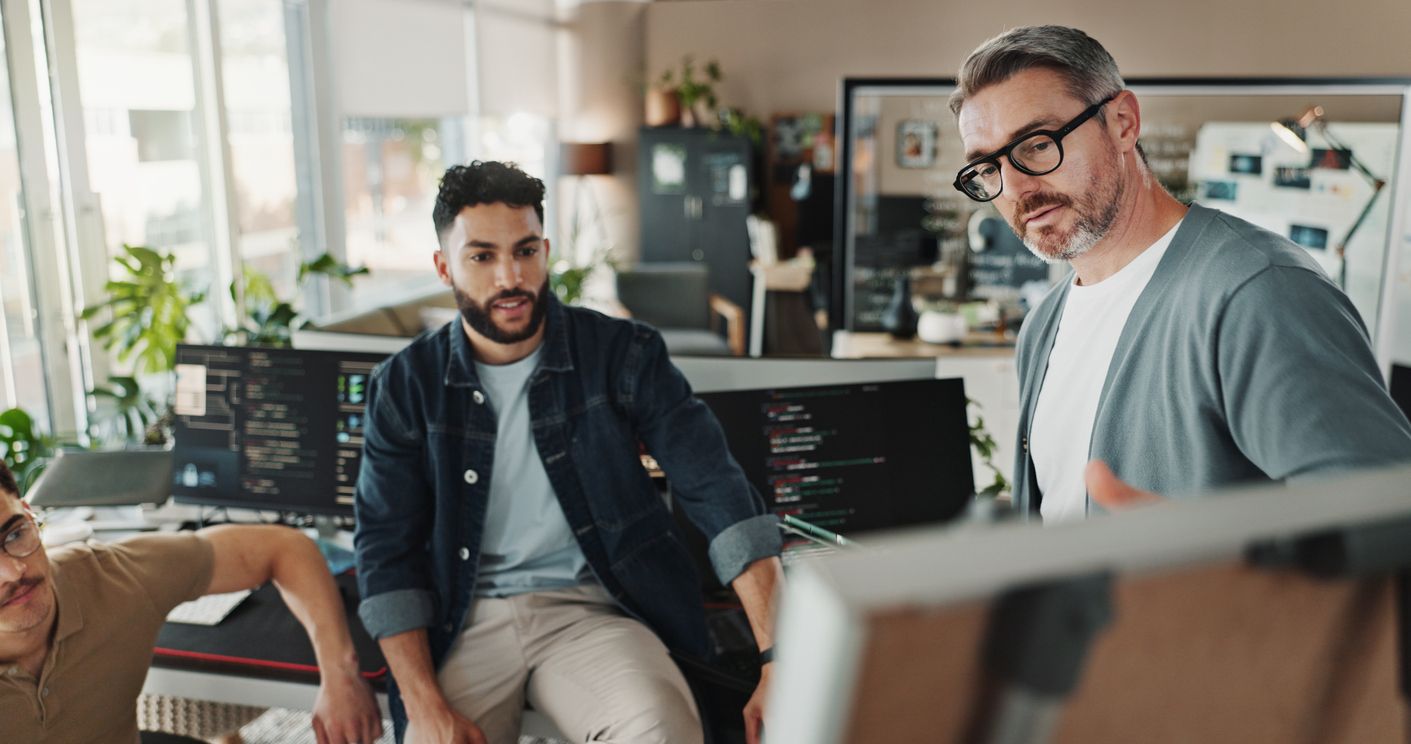 Three men sit around a computer monitor discussing computer programming.