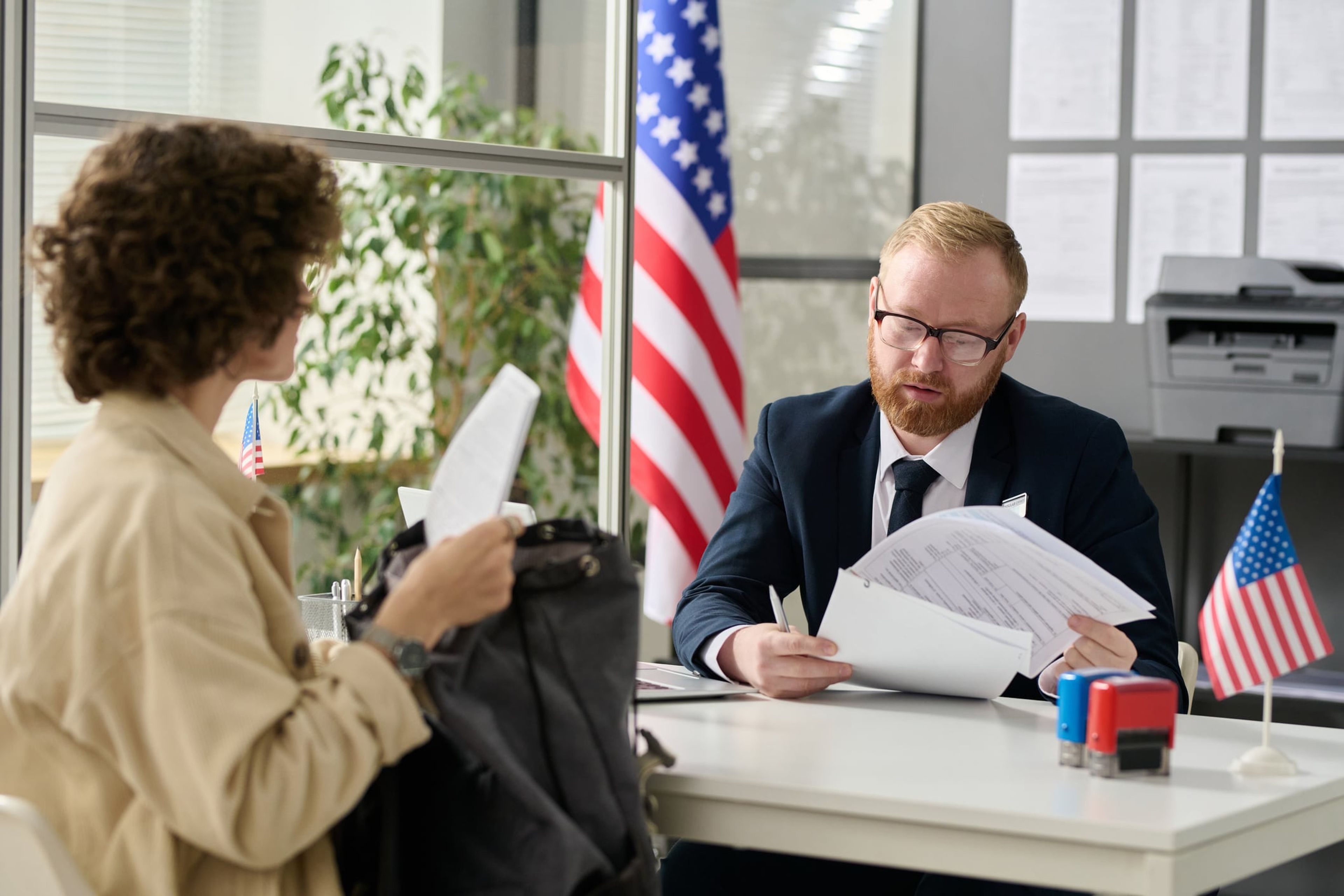 A young woman is seated at a desk in a U.S. immigration office while a seated man in a suit reviews her B-1/B-2 visa documents.