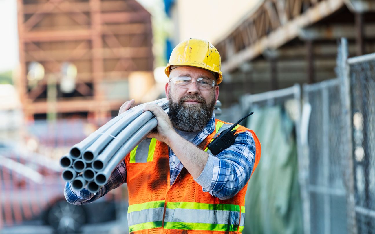 A male construction worker in a hard hat, protective glasses, and vest carrying a bundle of long pipes.
