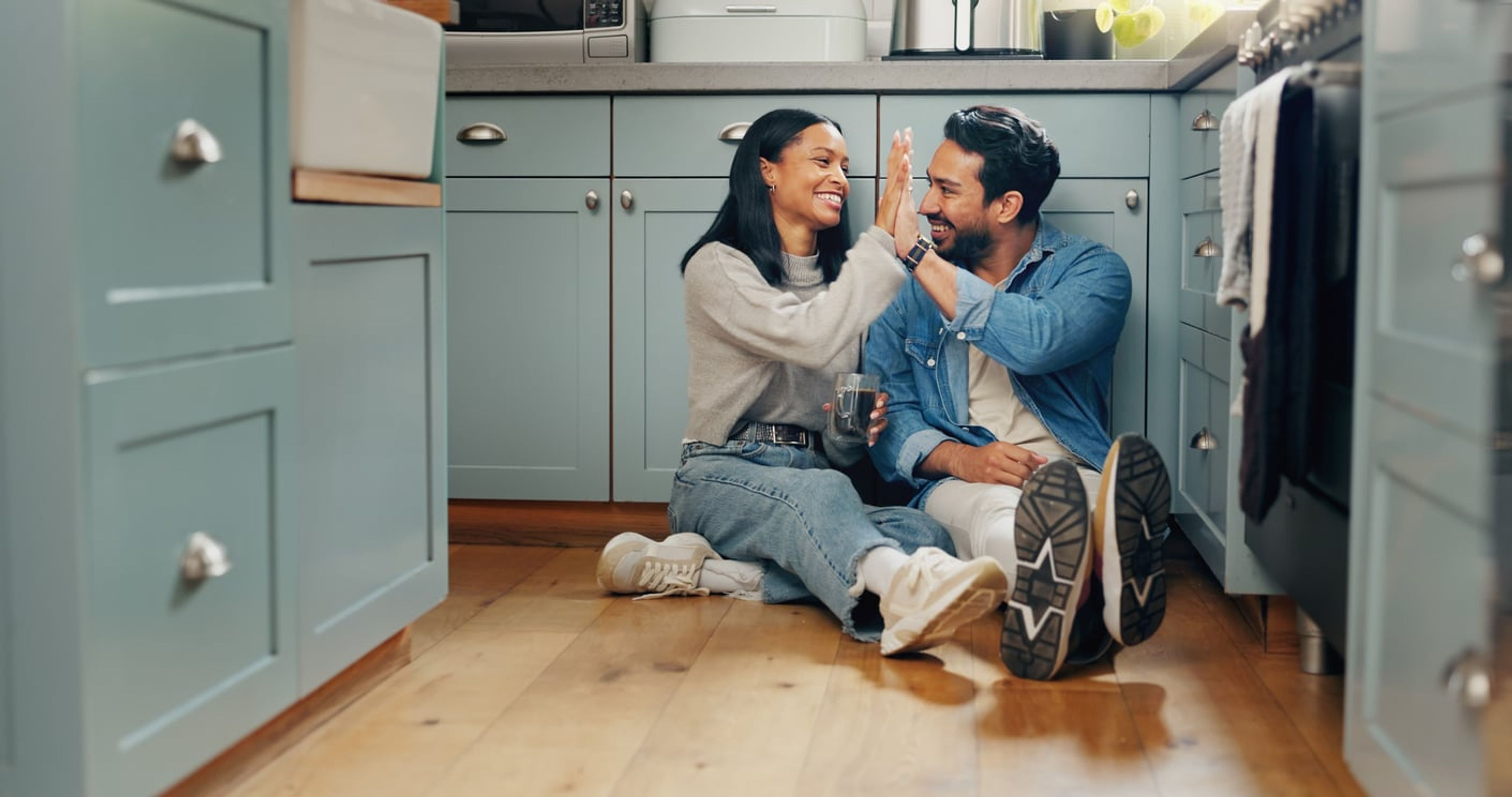 Happy married couple sitting on the floor of their kitchen clasping hands together.