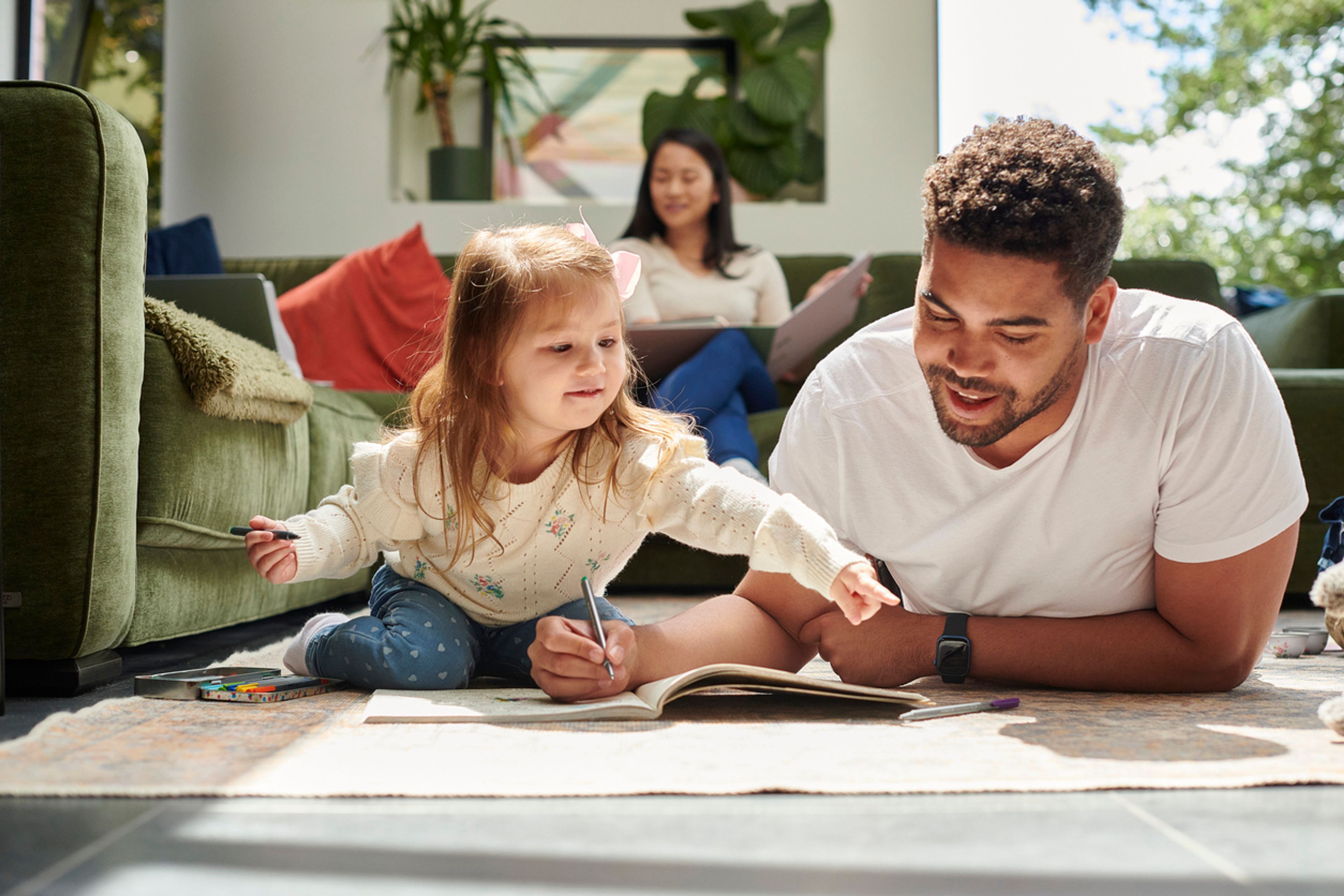 A man lays on the floor coloring with his daughter, while his spouse sits on the couch inthe background.