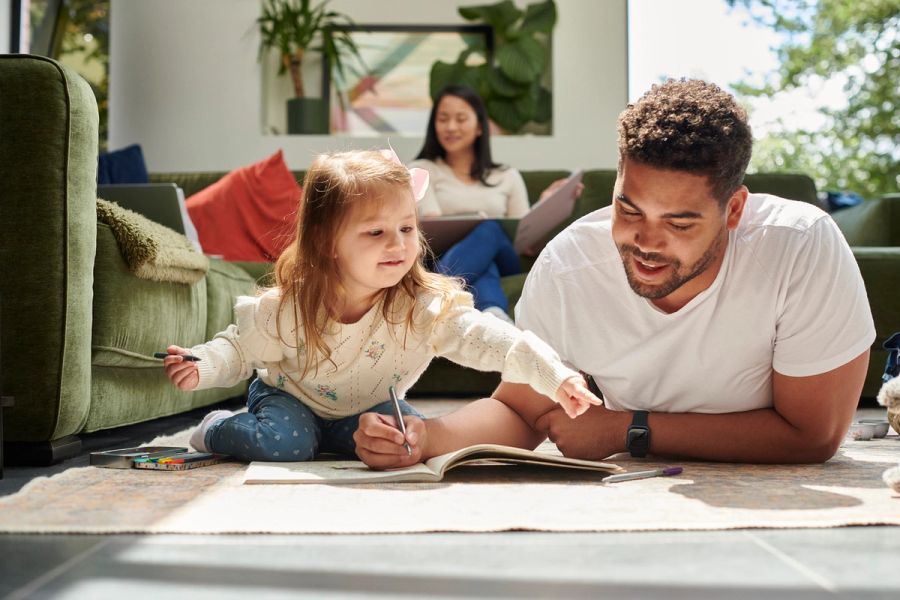 A man lays on the floor coloring with his daughter, while his spouse sits on the couch inthe background.