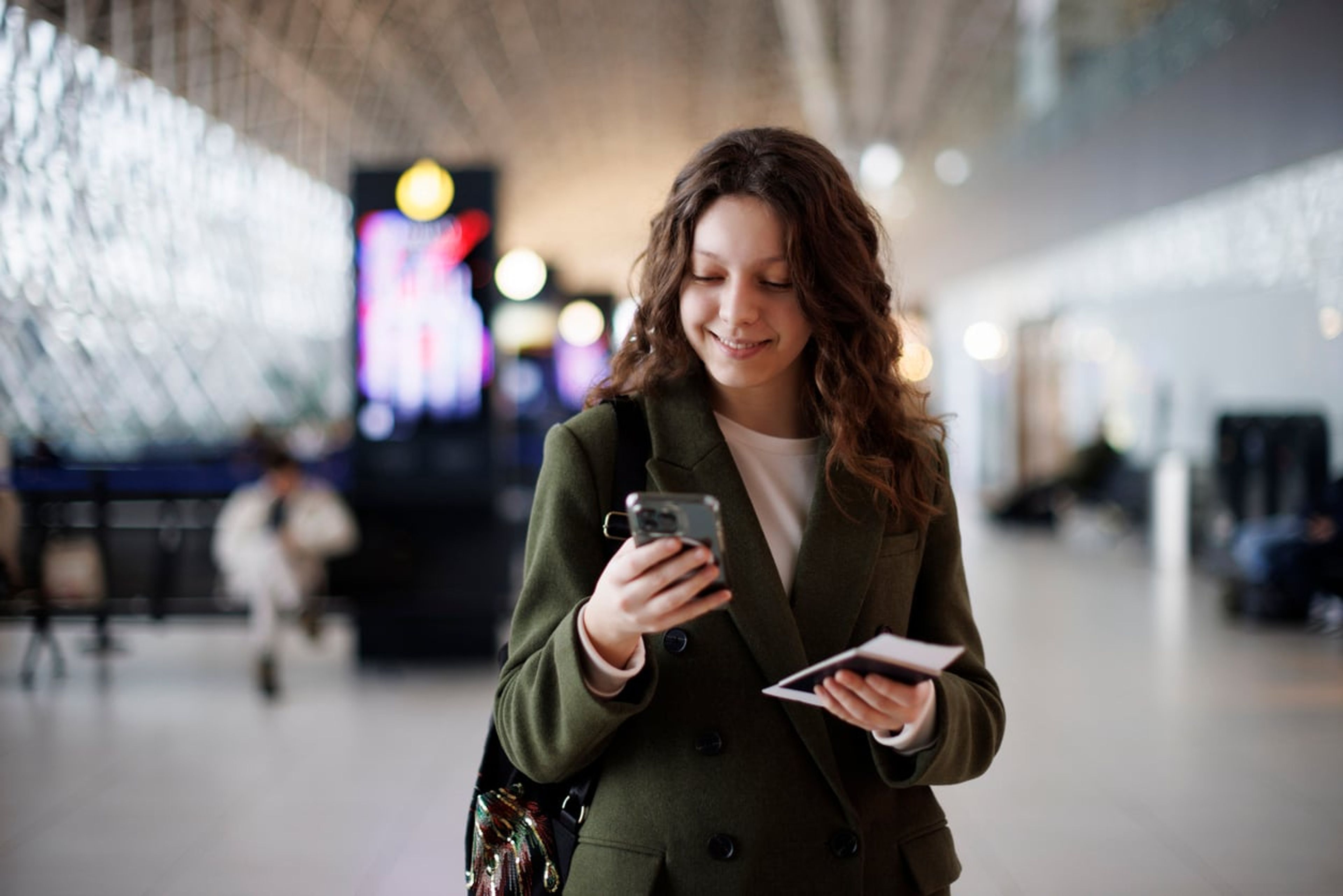 A young checks her phone at the airport while holding her passport wih boarding pass and Form I-551 stamp.