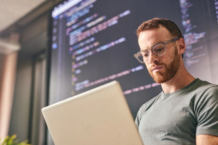 Software developer concentrating on his laptop, with lines of code displayed on a large screen in the background.