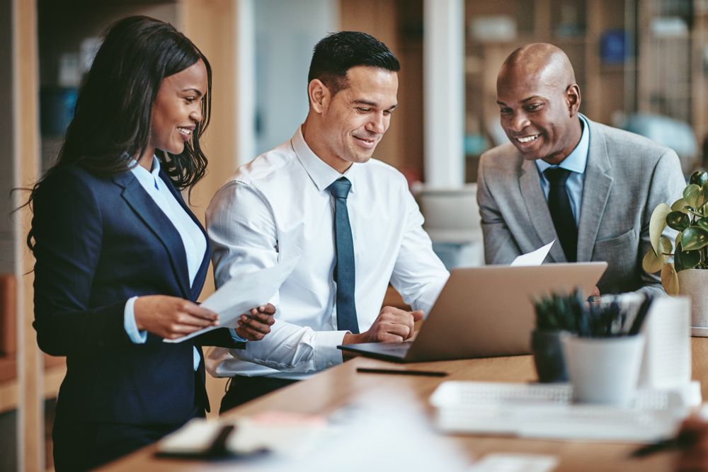 Three workers in a modern office looking at a laptop screen together.