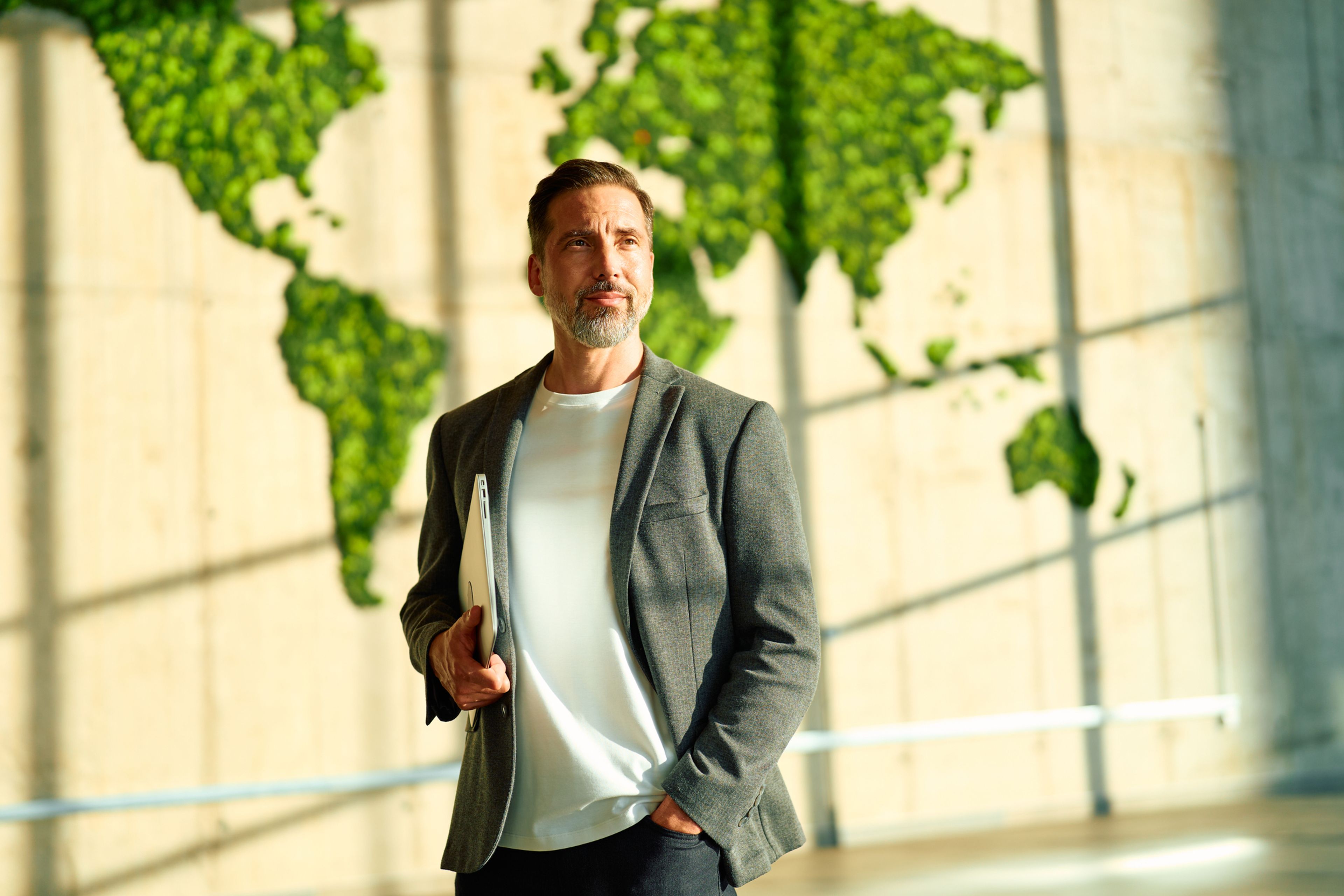 Professional executive standing in a modern office with a green moss world map on a wall, holding a digital notebook and contemplating global business strategies