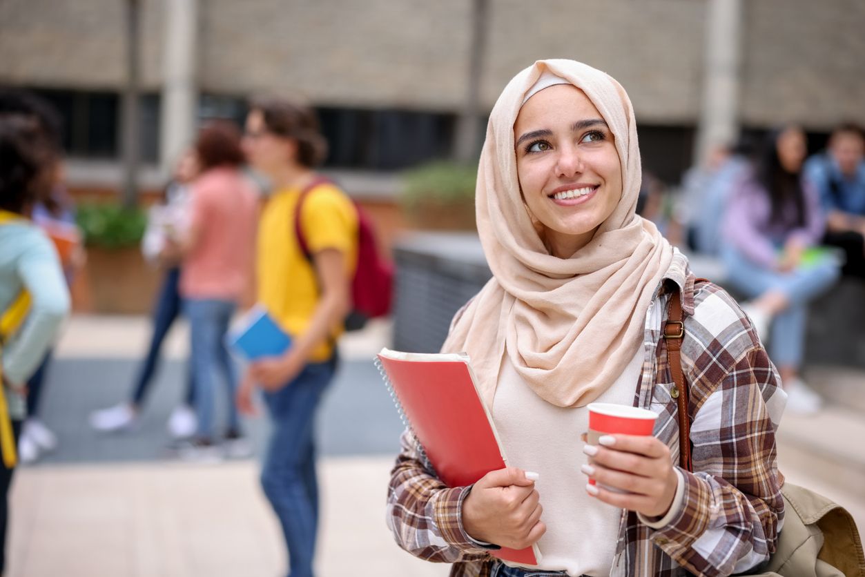 A young female student holding a notebook and cup smiles and looks up thoughtfully.