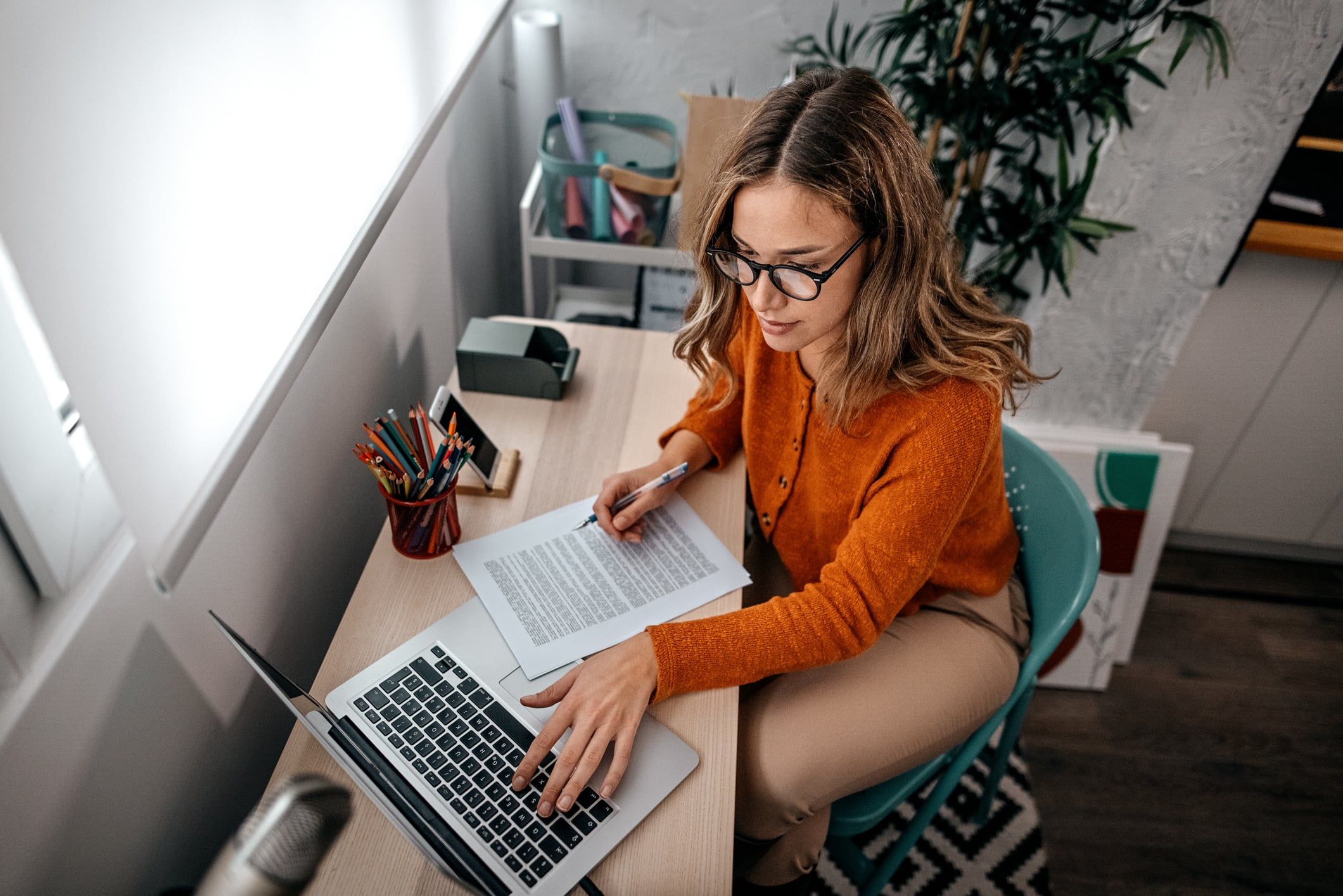A woman with glasses types on a laptop while while looking at a handwritten copy of an O-1 recommendation letter.
