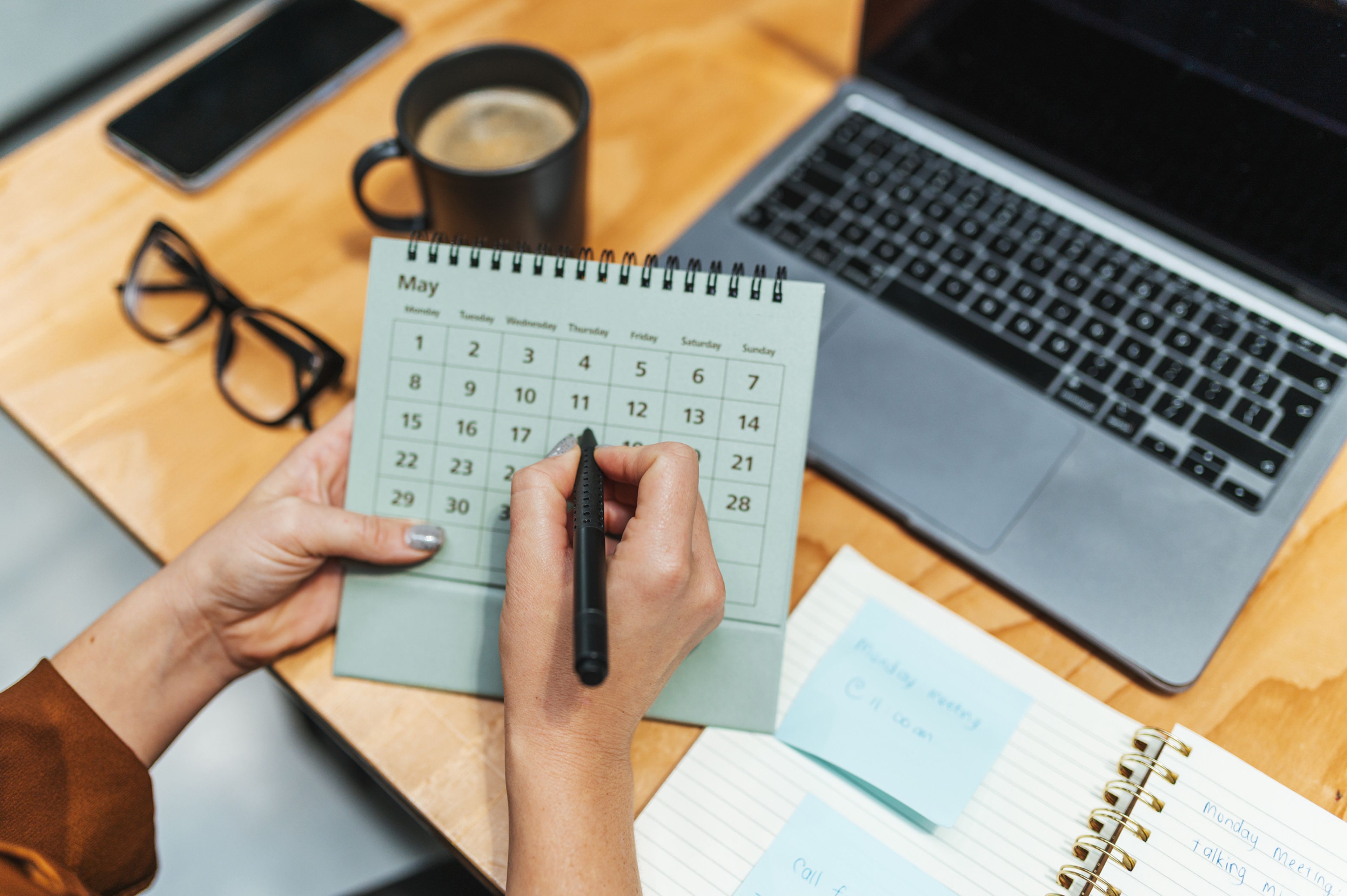 Person marking a date on a desk calendar next to a laptop.