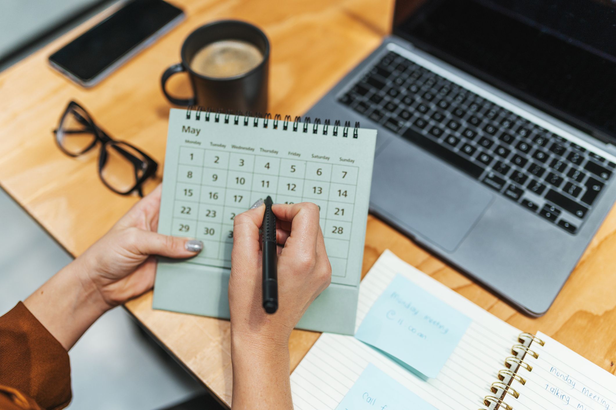 Person marking a date on a desk calendar next to a laptop.