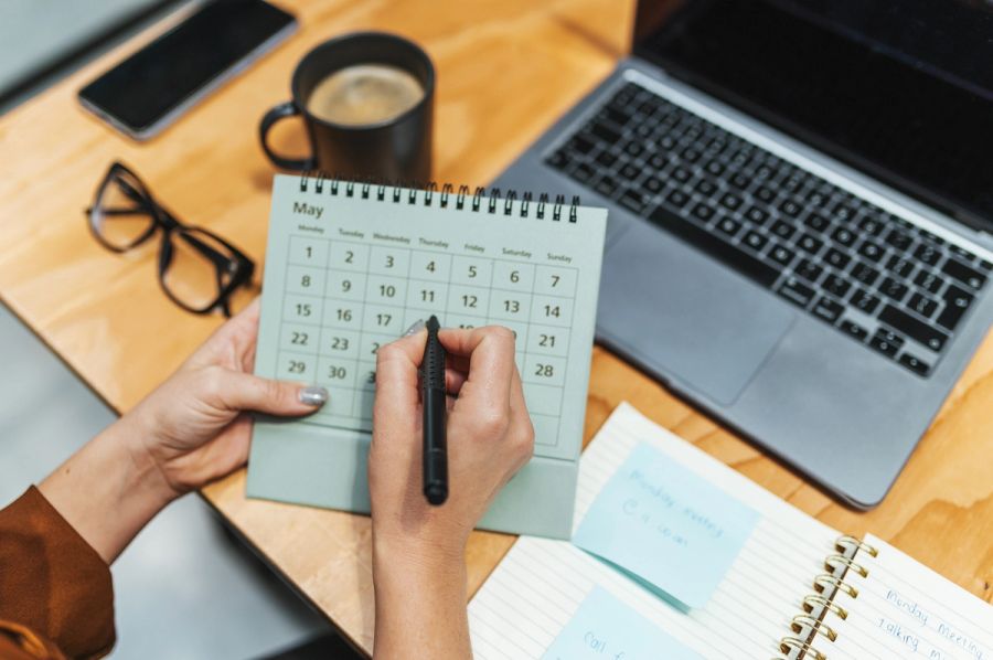 Person marking a date on a desk calendar next to a laptop.