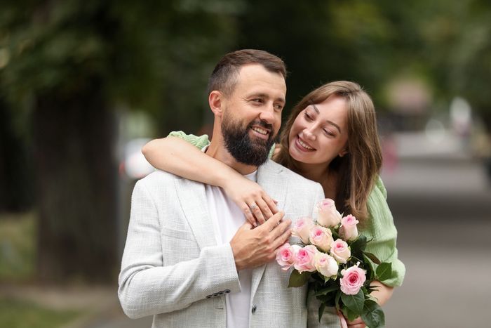 A man and a woman posing in an engagement photo displaying their rings and holding flowers
