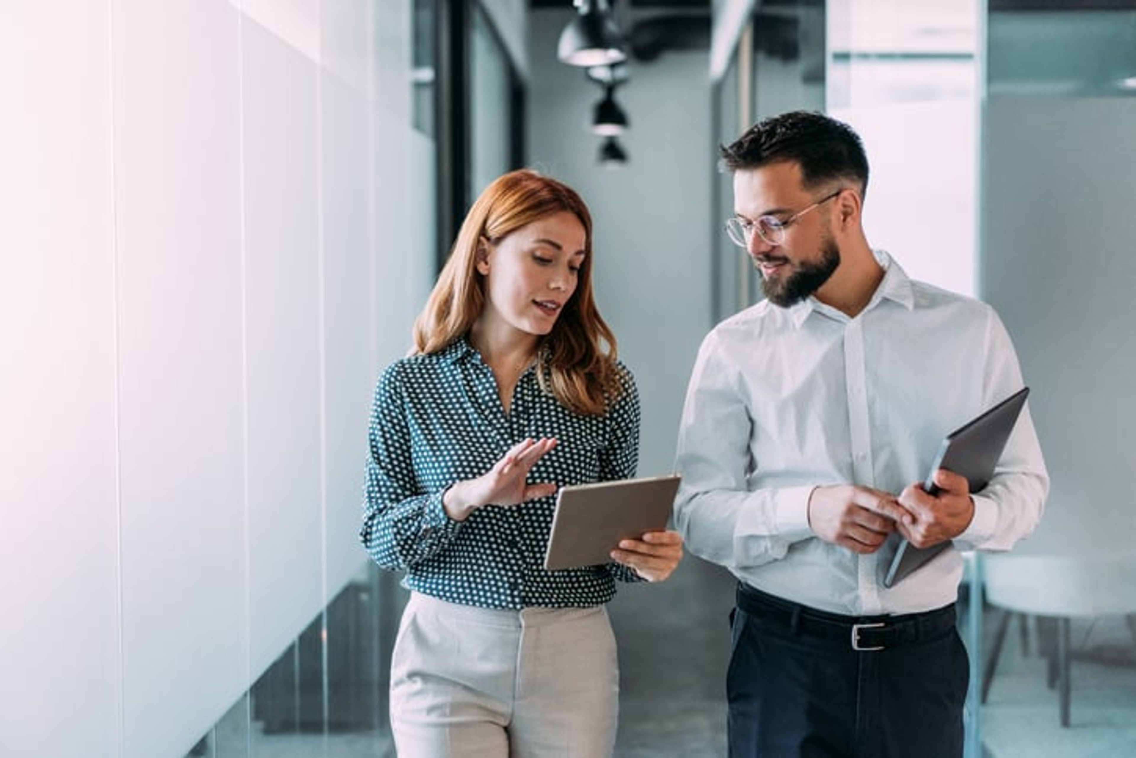 A man and woman are walking down an office hallway while discussing business strategy.