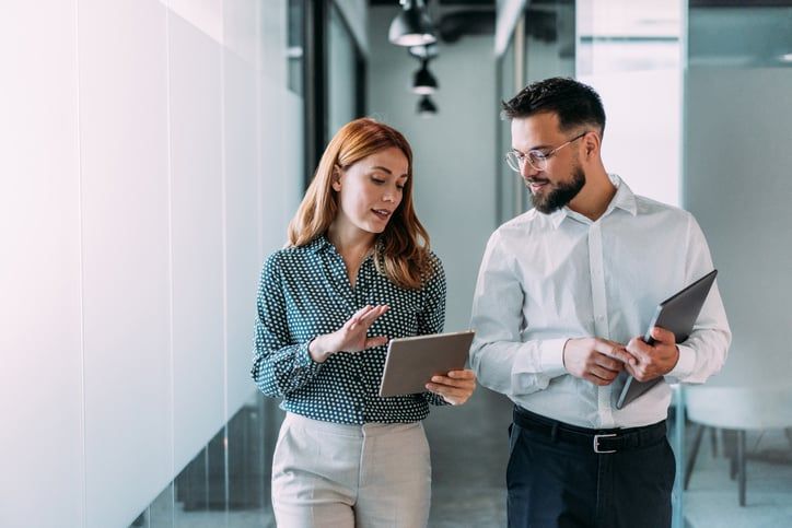 A man and woman are walking down an office hallway while discussing business strategy.