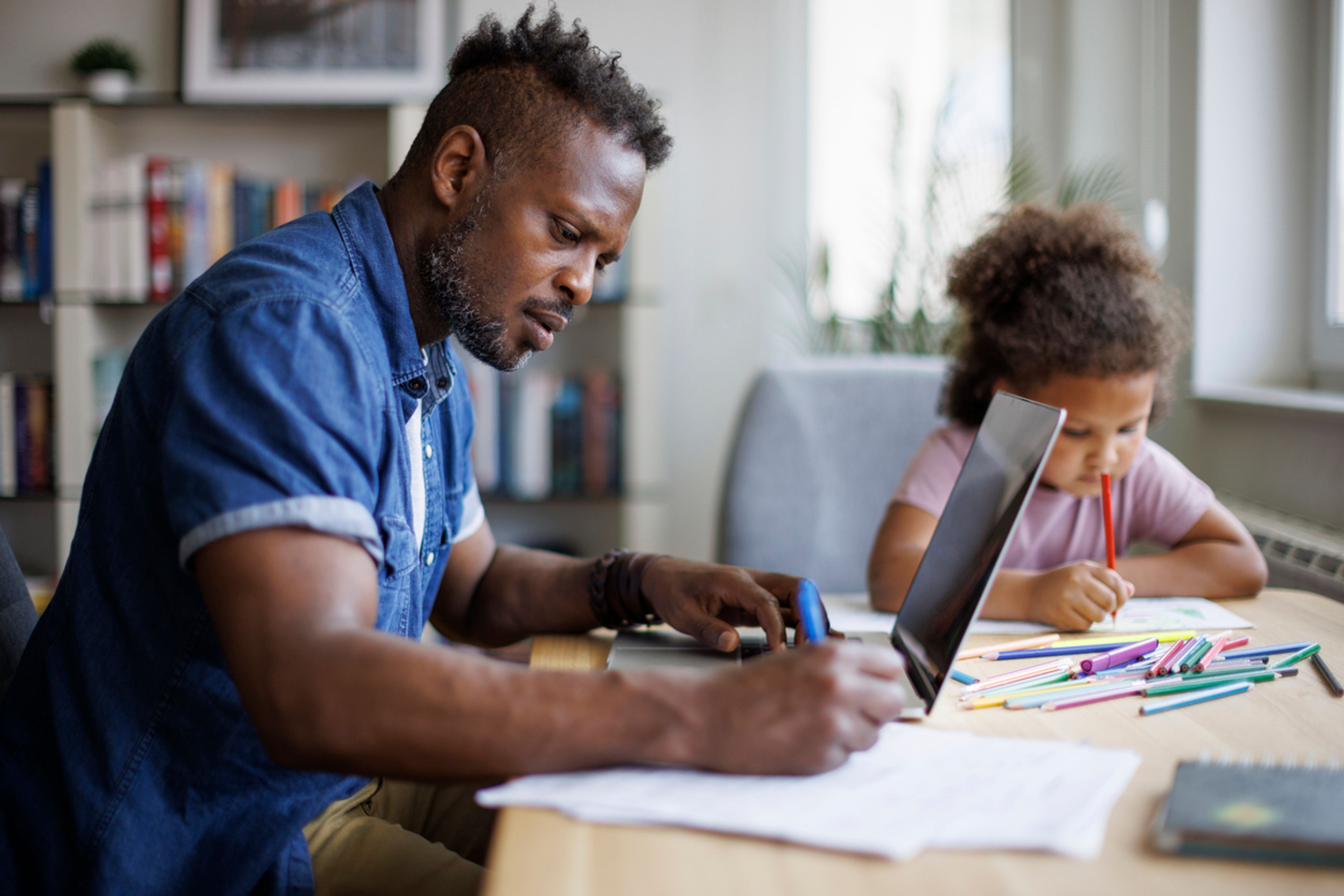 A seated man studies with a laptop and notebook while his daughter sits next to him with a coloring book.