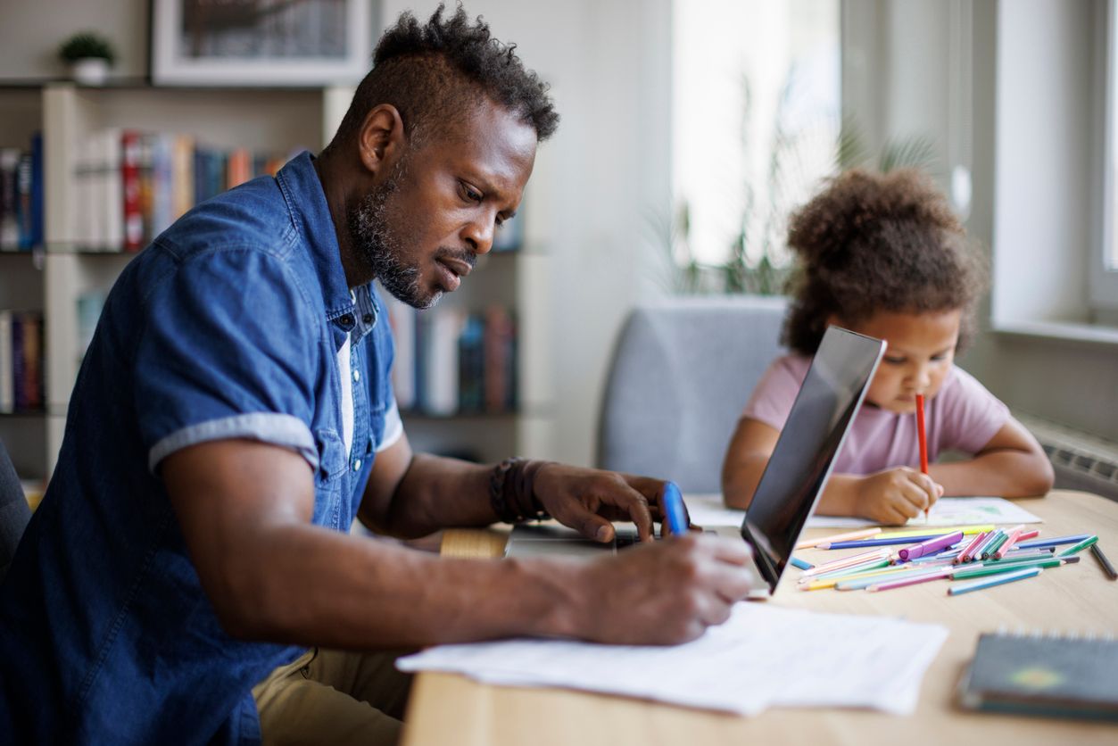 A seated man studies with a laptop and notebook while his daughter sits next to him with a coloring book.