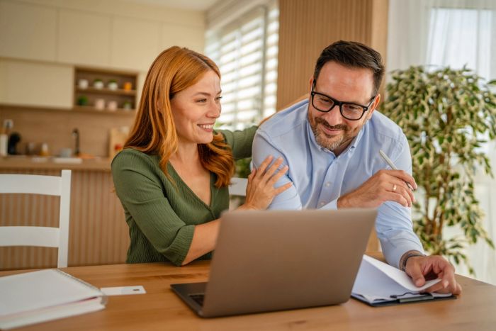 Happy couple working from home, looking at something on a laptop in their kitchen.
