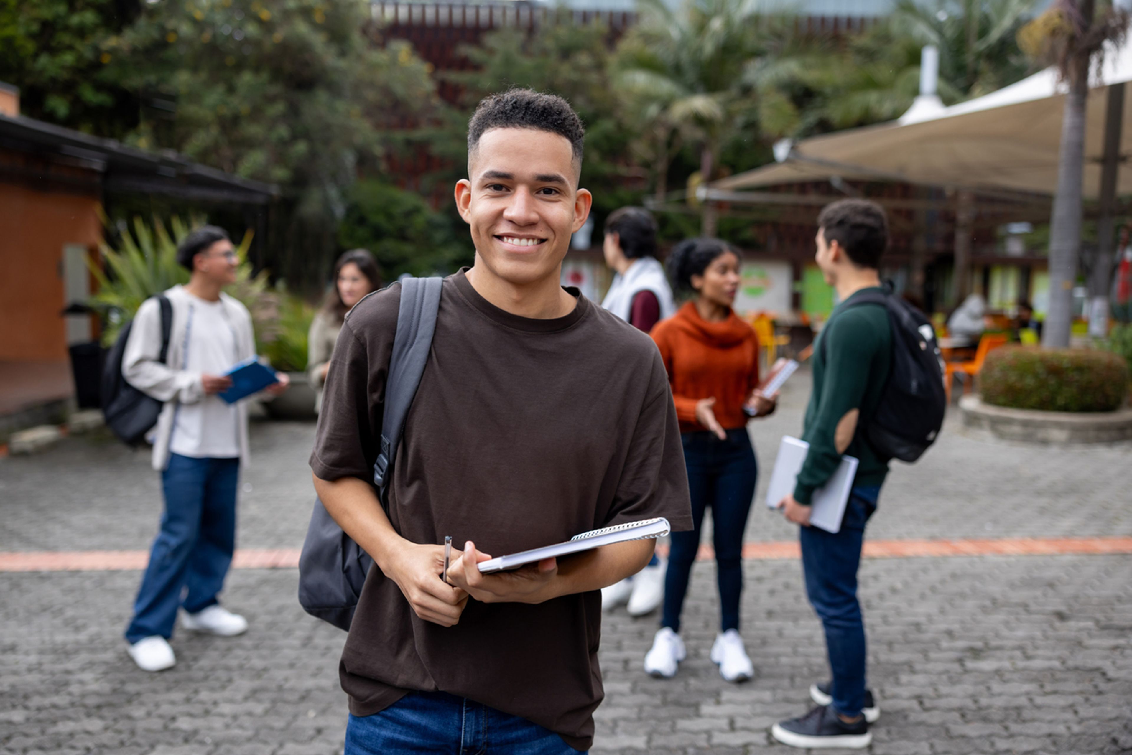 College student on campus with other students, holding a notebook.