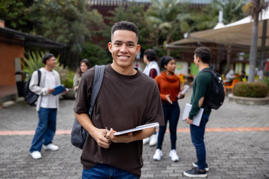 College student on campus with other students, holding a notebook.