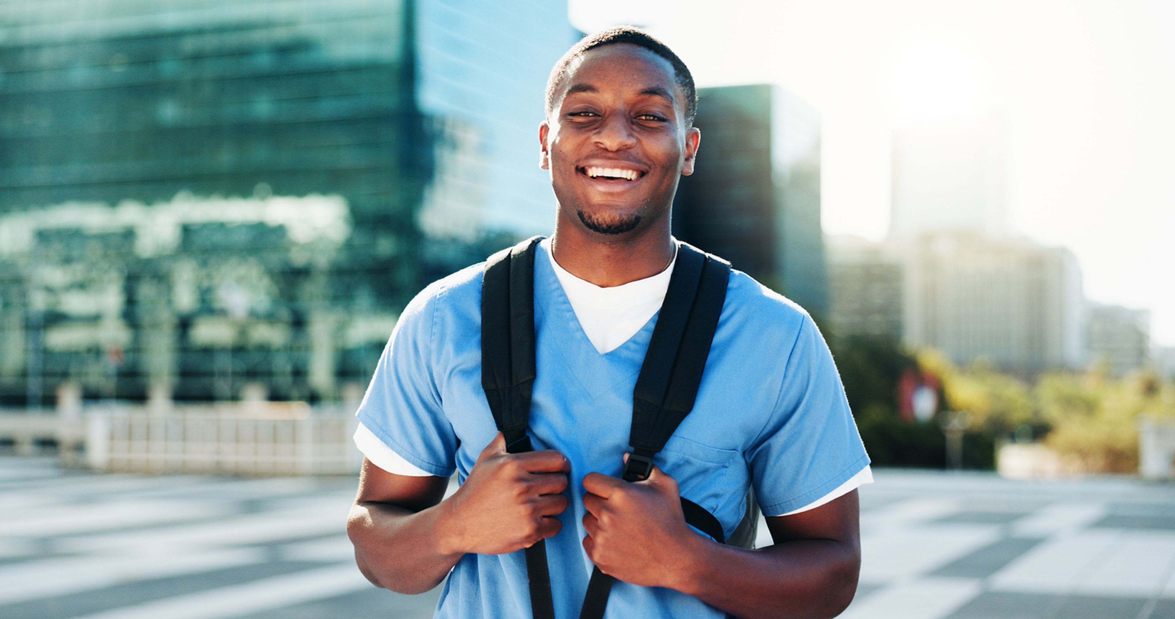 A young man in scrubs and a backpack stands facing the camera and smiling.
