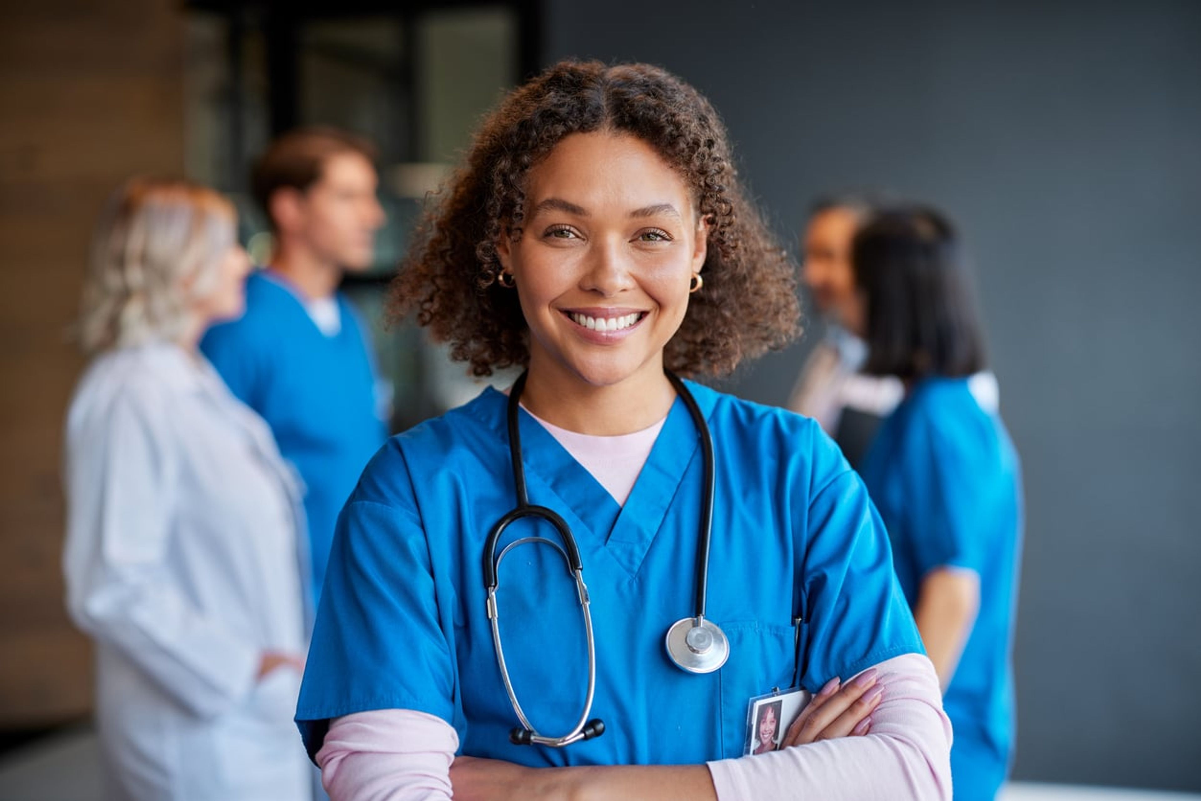 A female nurse in blue scrubs with crossed arms smiles at the camera.
