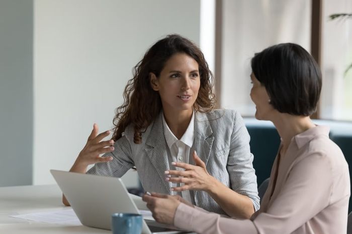 Two business women sit at desk discussing project details.