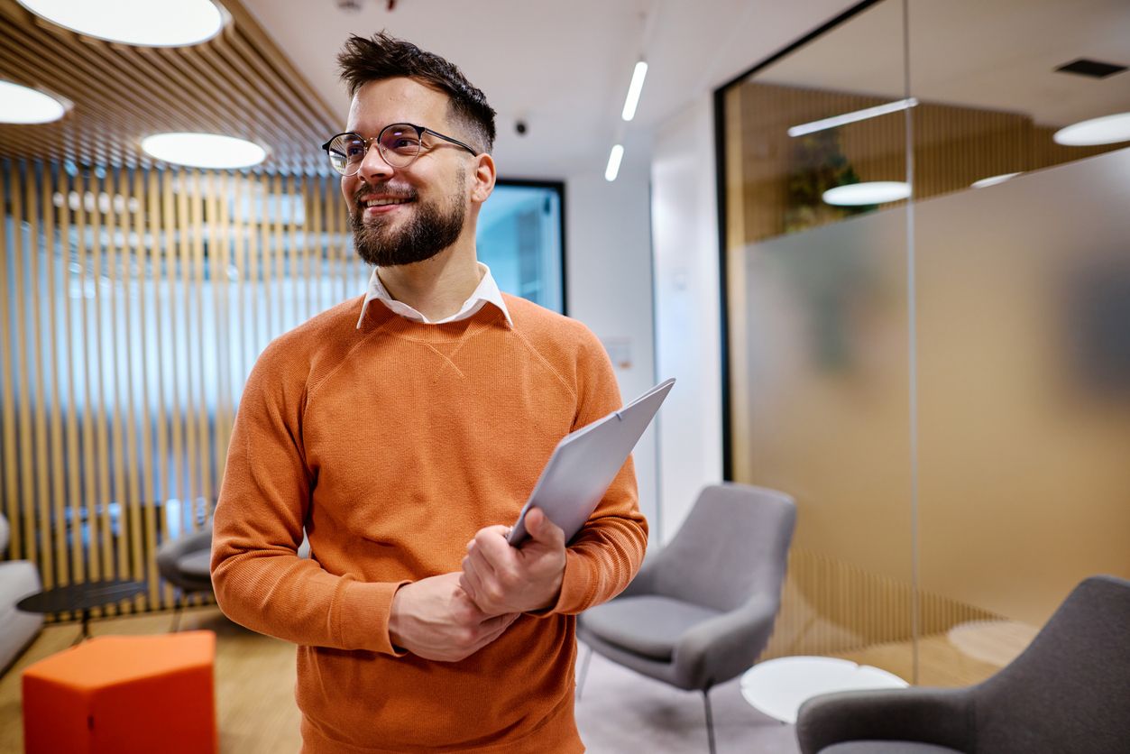 Young businessman holding a tablet and smiling in a modern office, looking away with a sense of confidence and ambition.