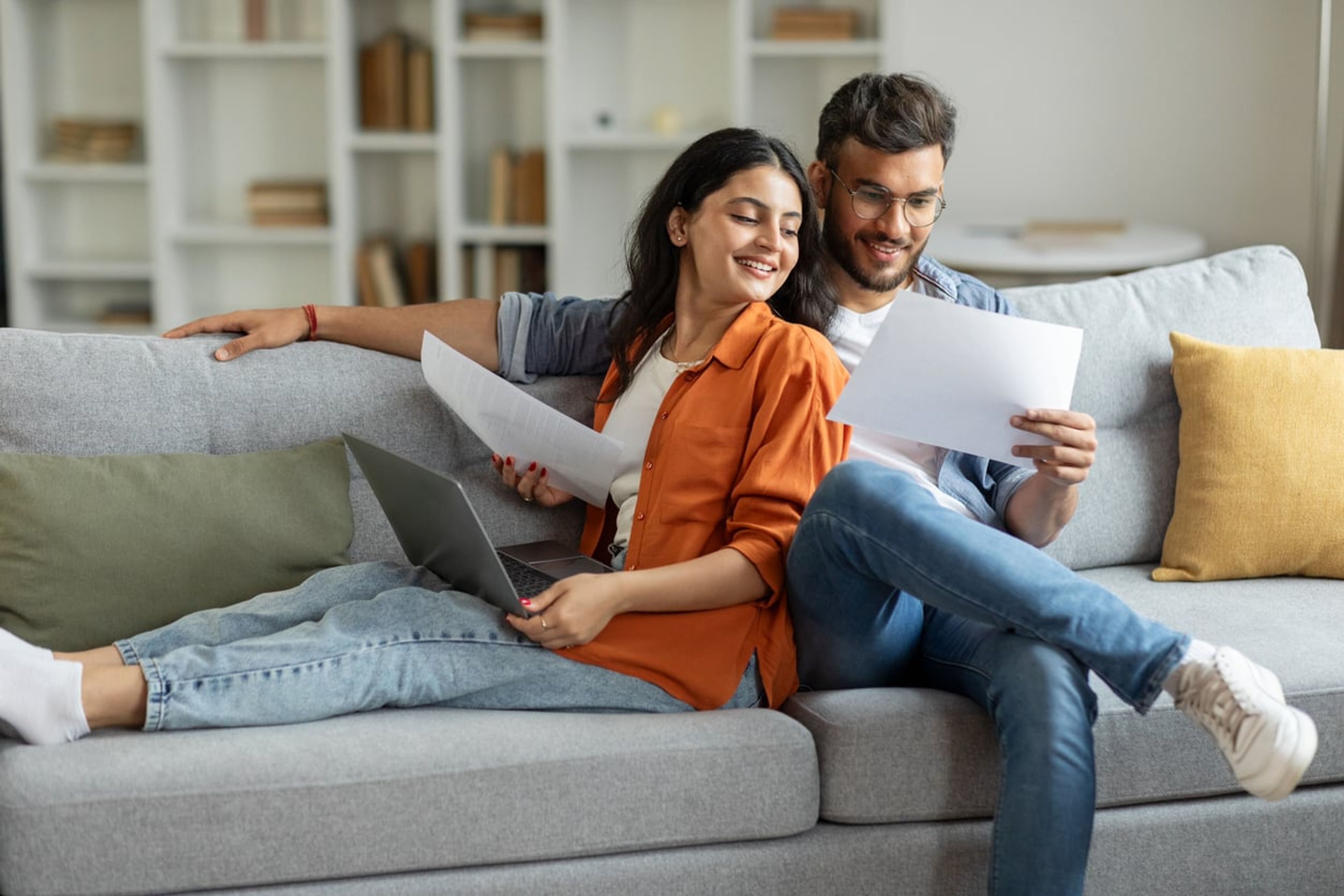 A married man and woman sit on a couch, smiling and looking at documents together.
