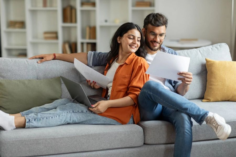 A married man and woman sit on a couch, smiling and looking at documents together.