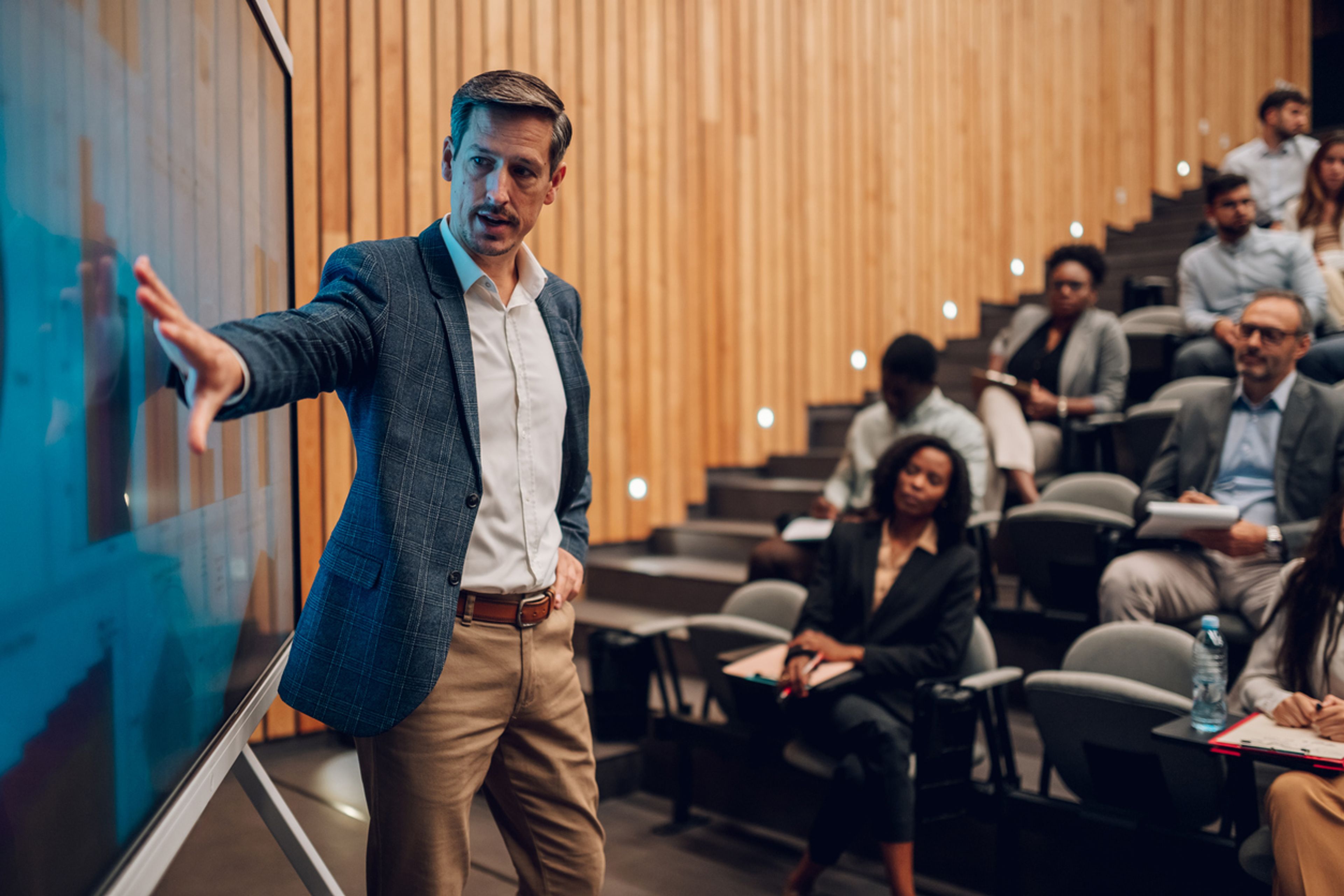 Professor in a university lecture hall gesturing to a board in front of seated students.