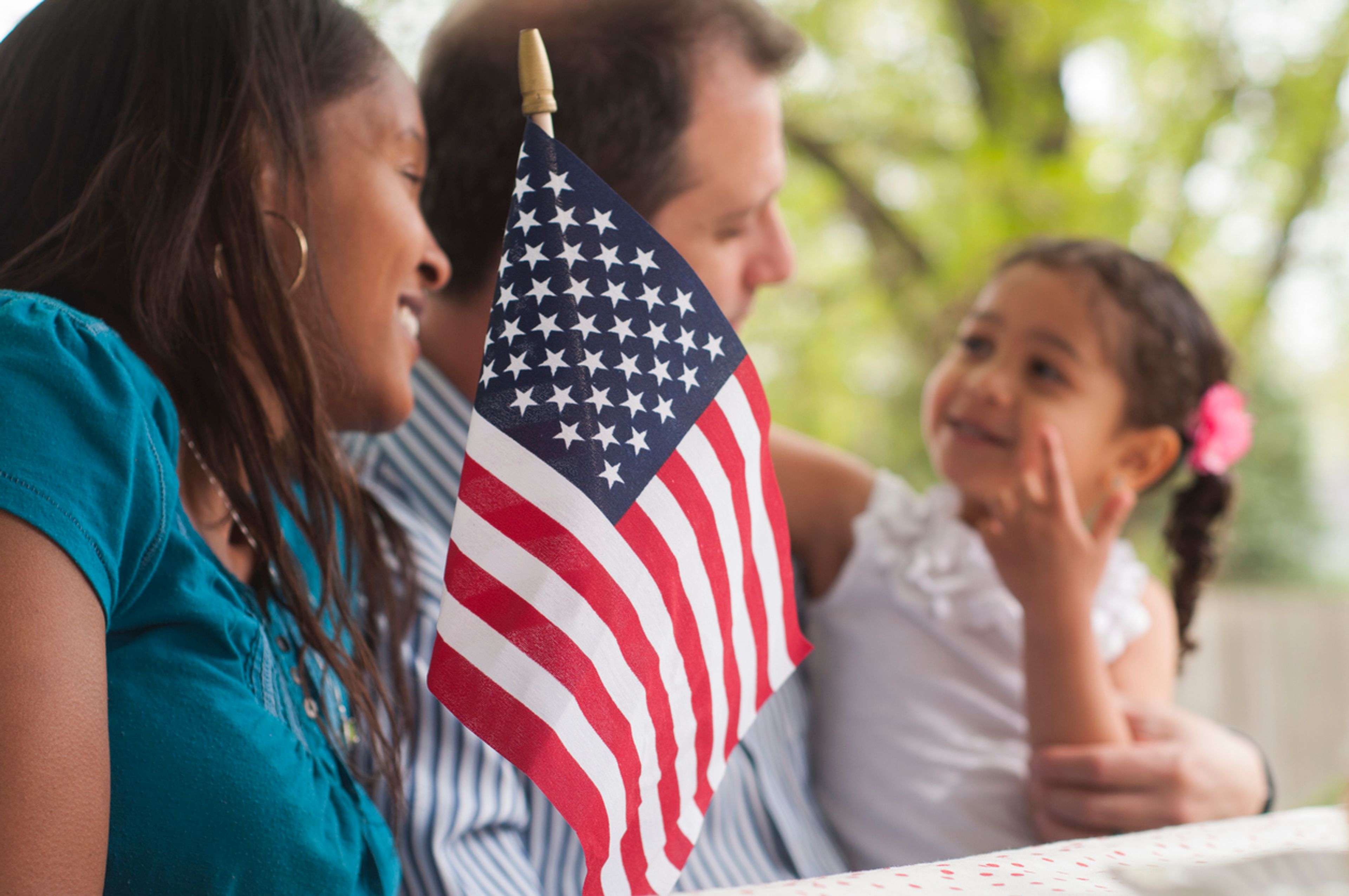 A husband and wife with their daughter sit at a park bench outside with an American flag placed in front of them.