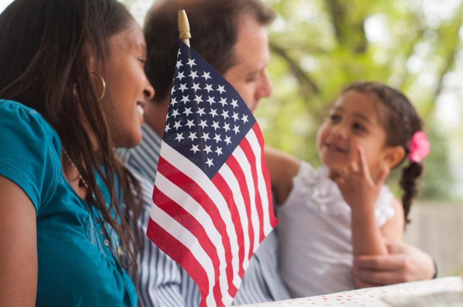 A husband and wife with their daughter sit at a park bench outside with an American flag placed in front of them.