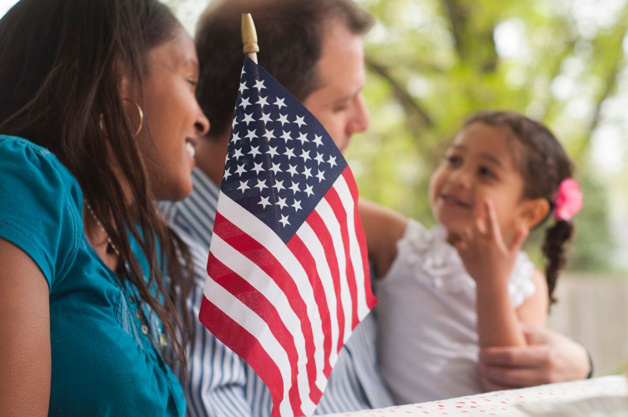 A husband and wife with their daughter sit at a park bench outside with an American flag placed in front of them.