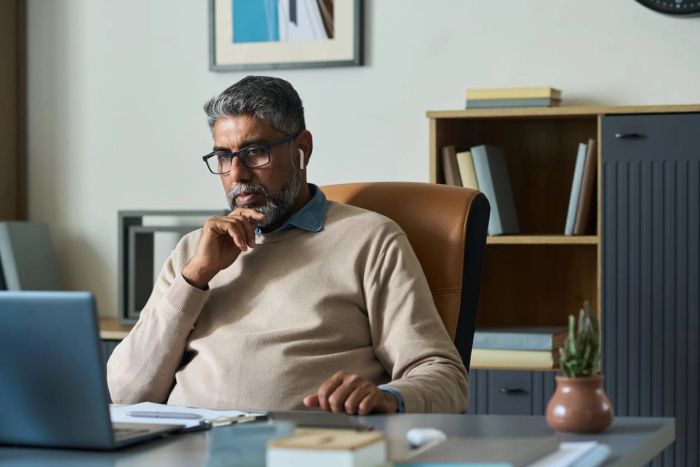 A middle-aged South Asian man with glasses and ear buds sits at a desk and looks at his laptop during an executive meeting.
