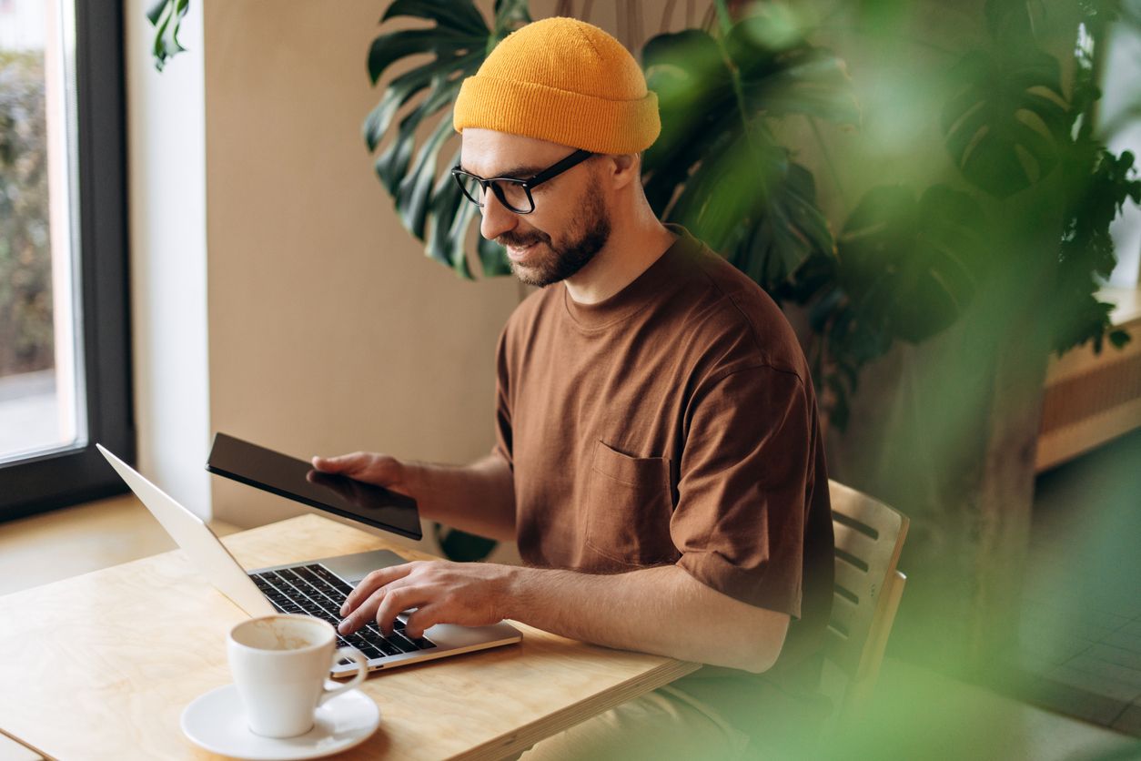 smiling man wearing yellow hat and eyeglasses holds tablet at table