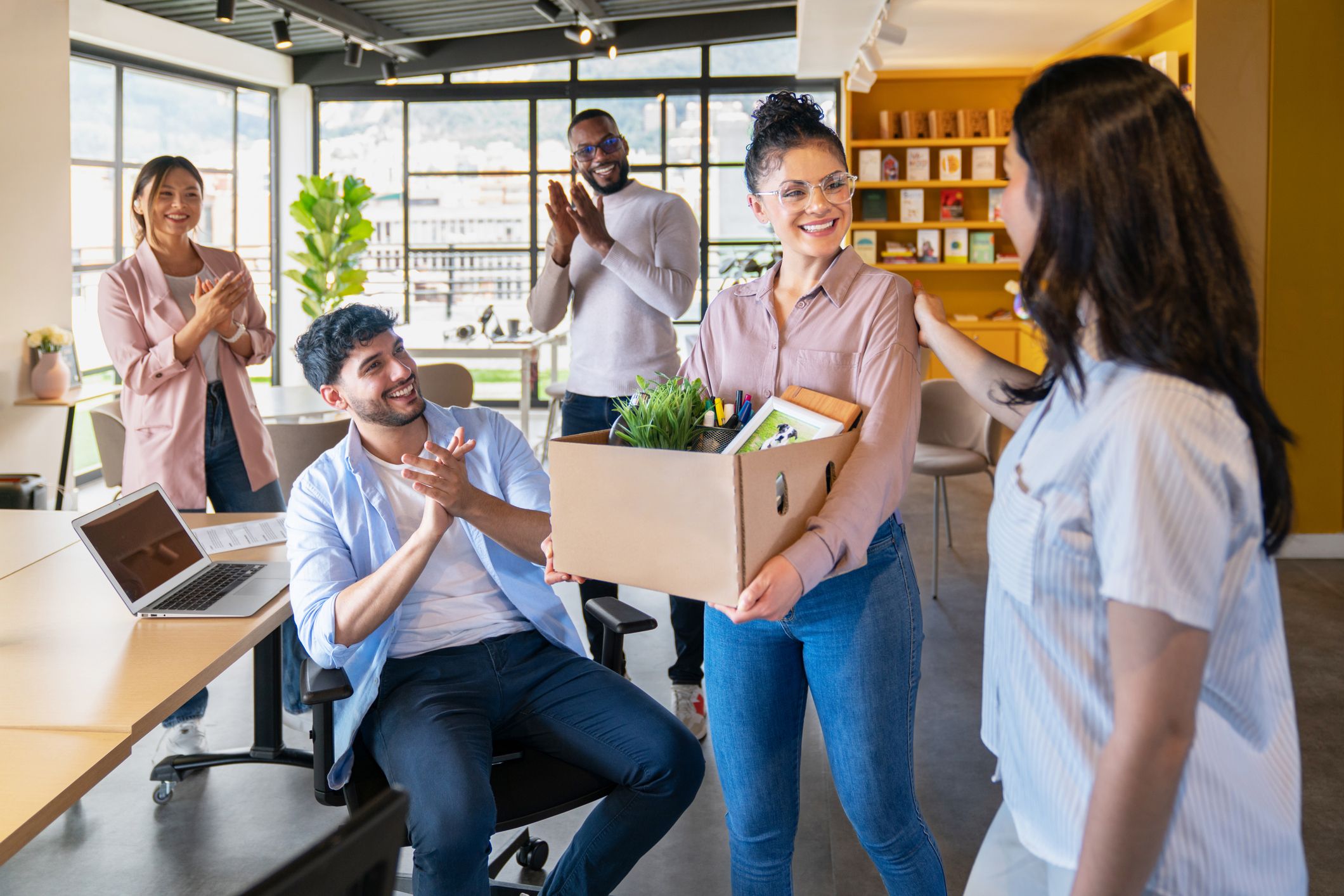 Smiling professional carrying a box of belongings into a new office as colleagues welcome her.