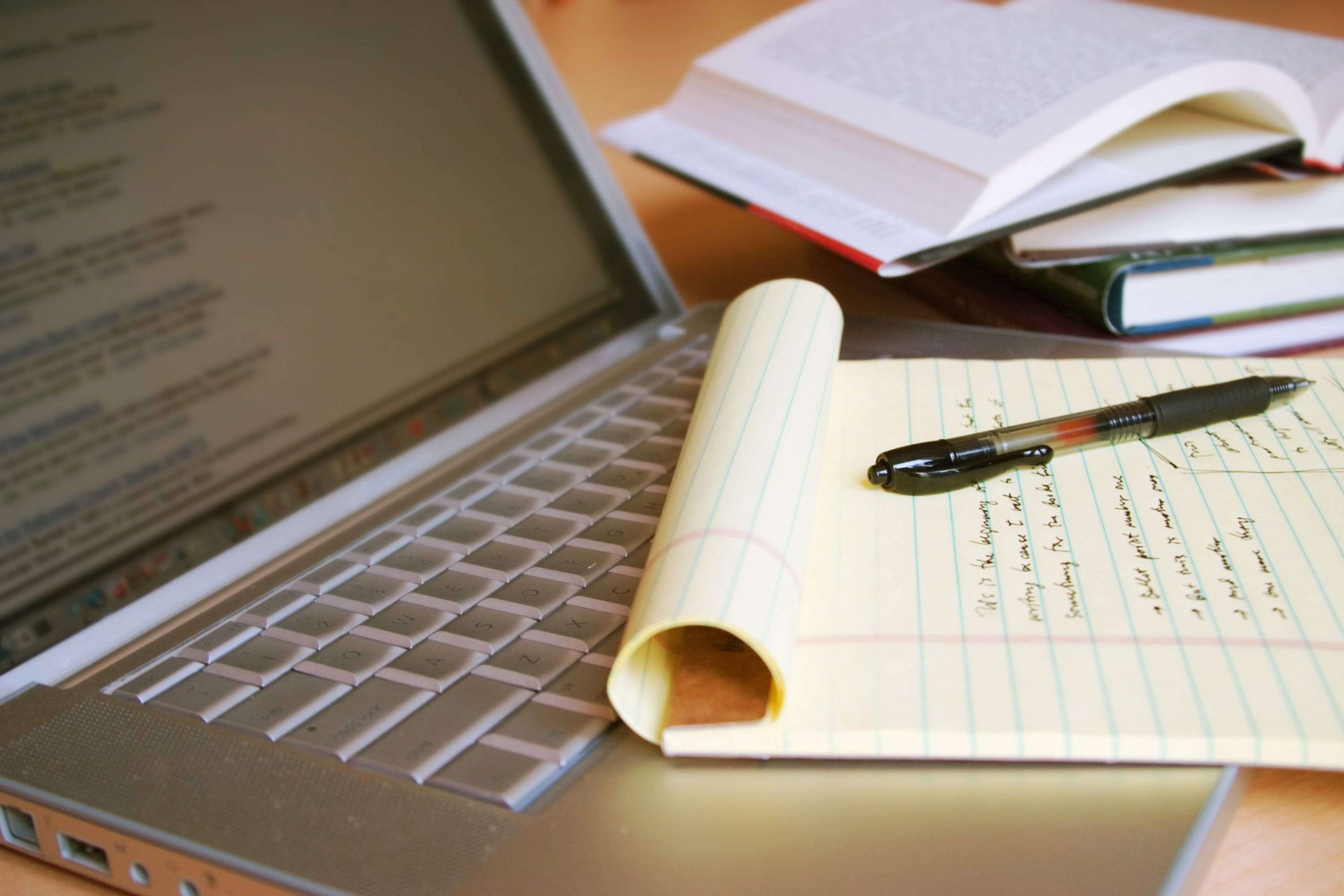 Closeup of a yellow legal pad and a pen resting on the keyboard of a laptop beside a pile of books