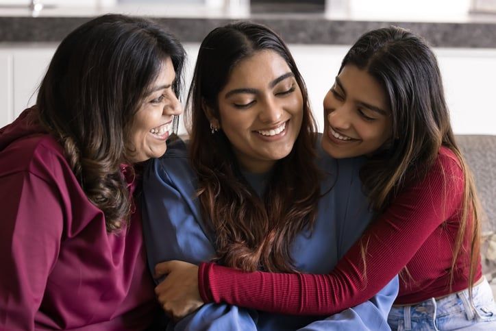 Three adult sisters sit smiling at each other and hugging.