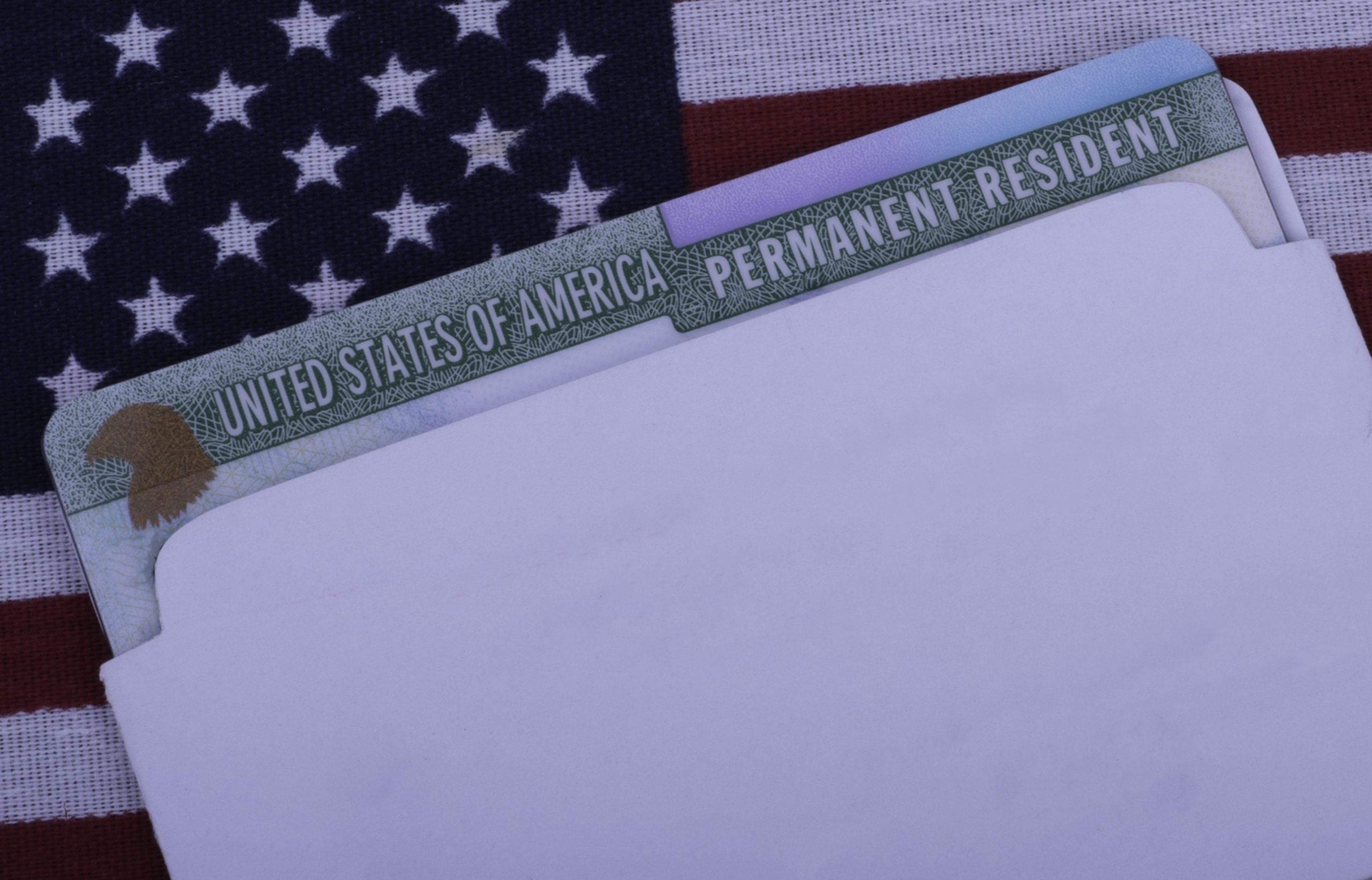 A Green card or permanent resident card is partially showing out of an envelope as it lays atop a U.S. flag.